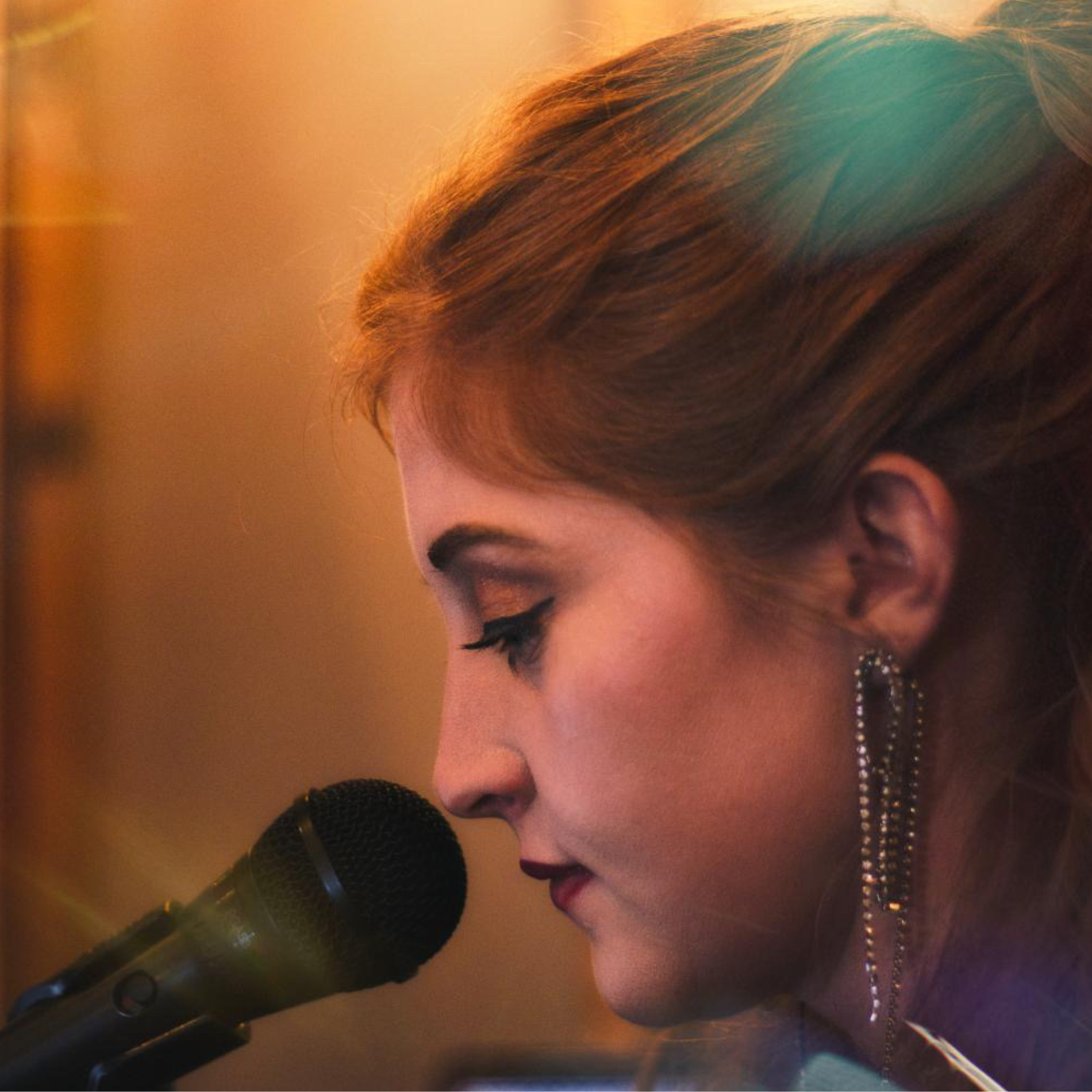 A woman singing into a microphone with her eyes closed, wearing long earrings and makeup, in a warm indoor setting.