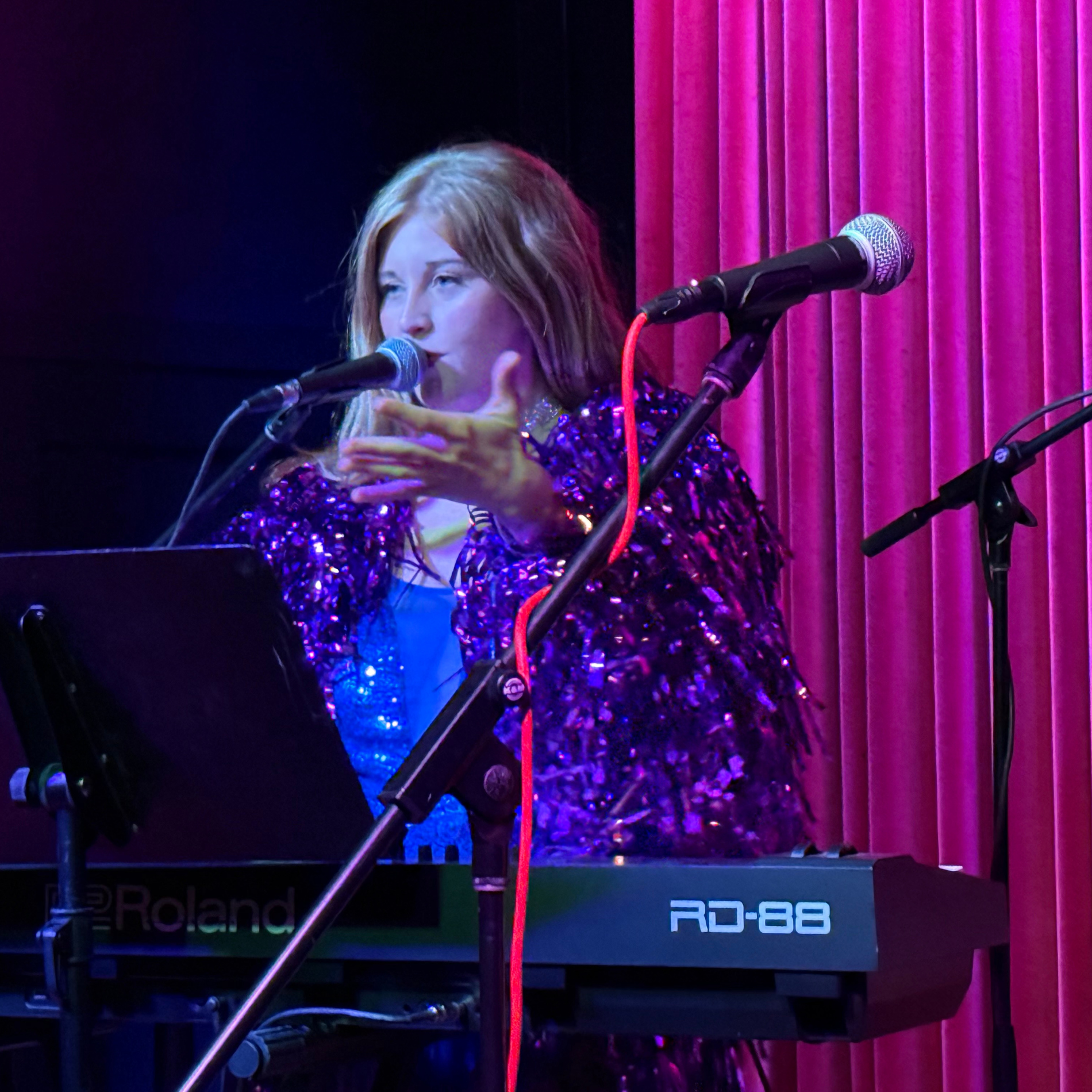 A woman with long blonde hair singing into a microphone while playing a Roland keyboard on stage, with a pink curtain background.