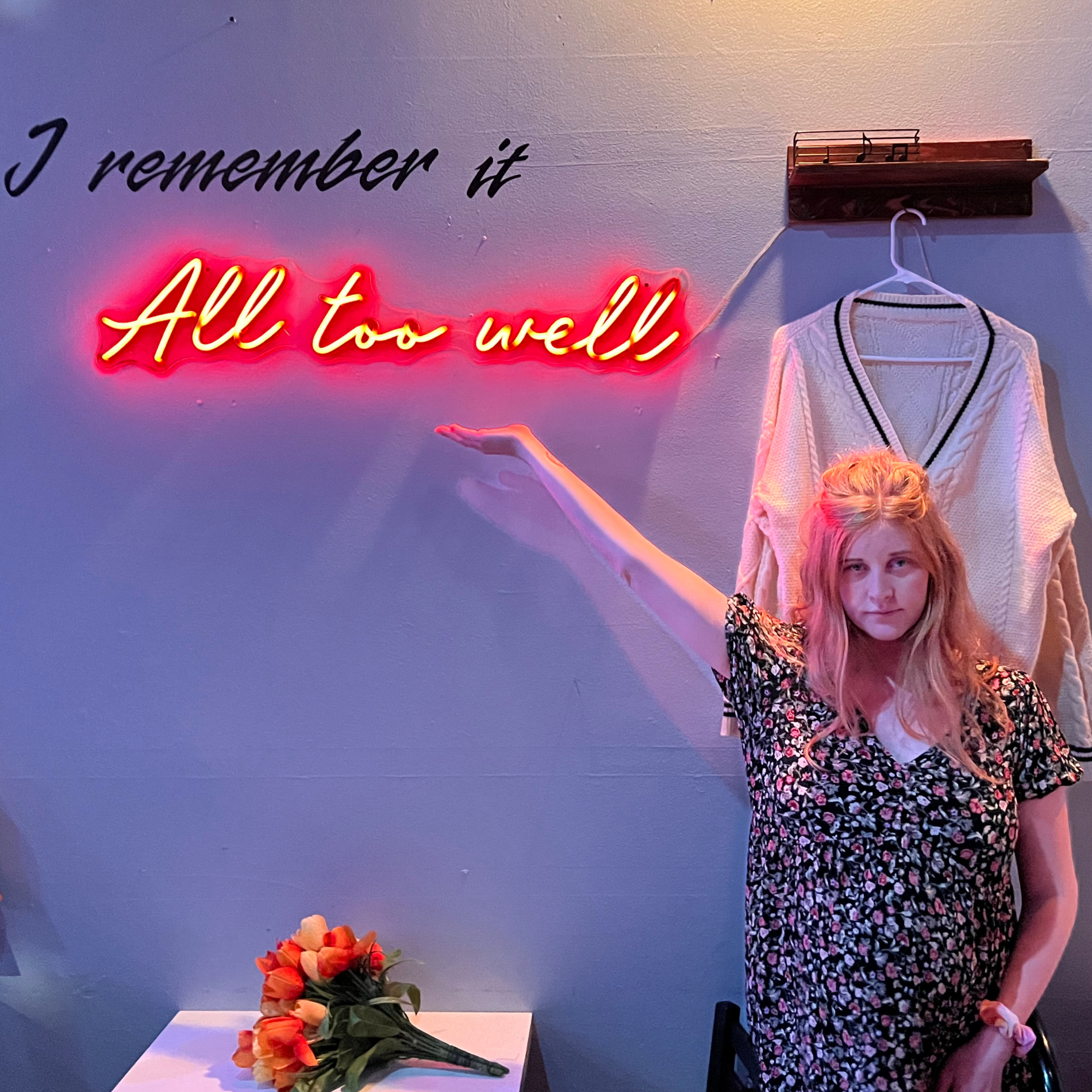 A woman with pink hair in a floral dress points to a neon sign on the wall that reads 'All too well' in cursive. There is a white sweater hanging on a hook behind her and a bouquet of orange and yellow tulips on a table in front of her.