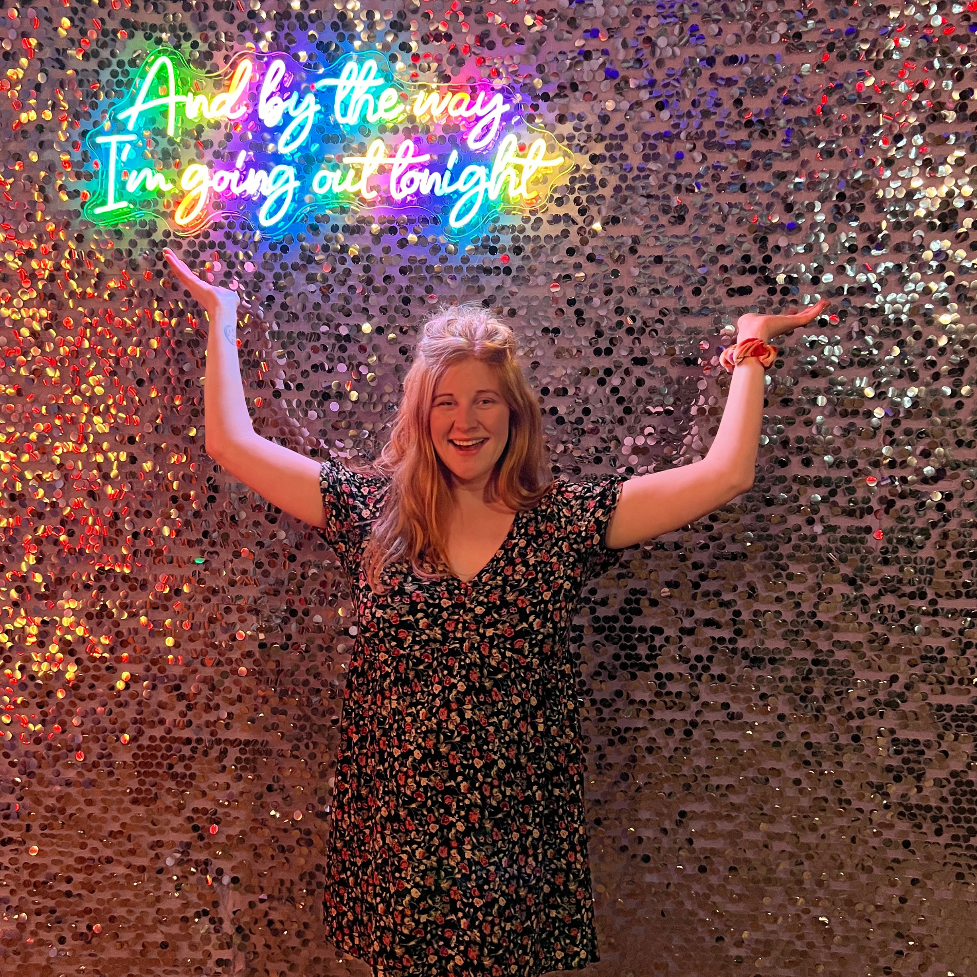 A woman with red hair, wearing a floral dress, smiling with arms raised, standing in front of a sequin wall and a colorful neon sign that says 'And by the way, I'm going out tonight.'