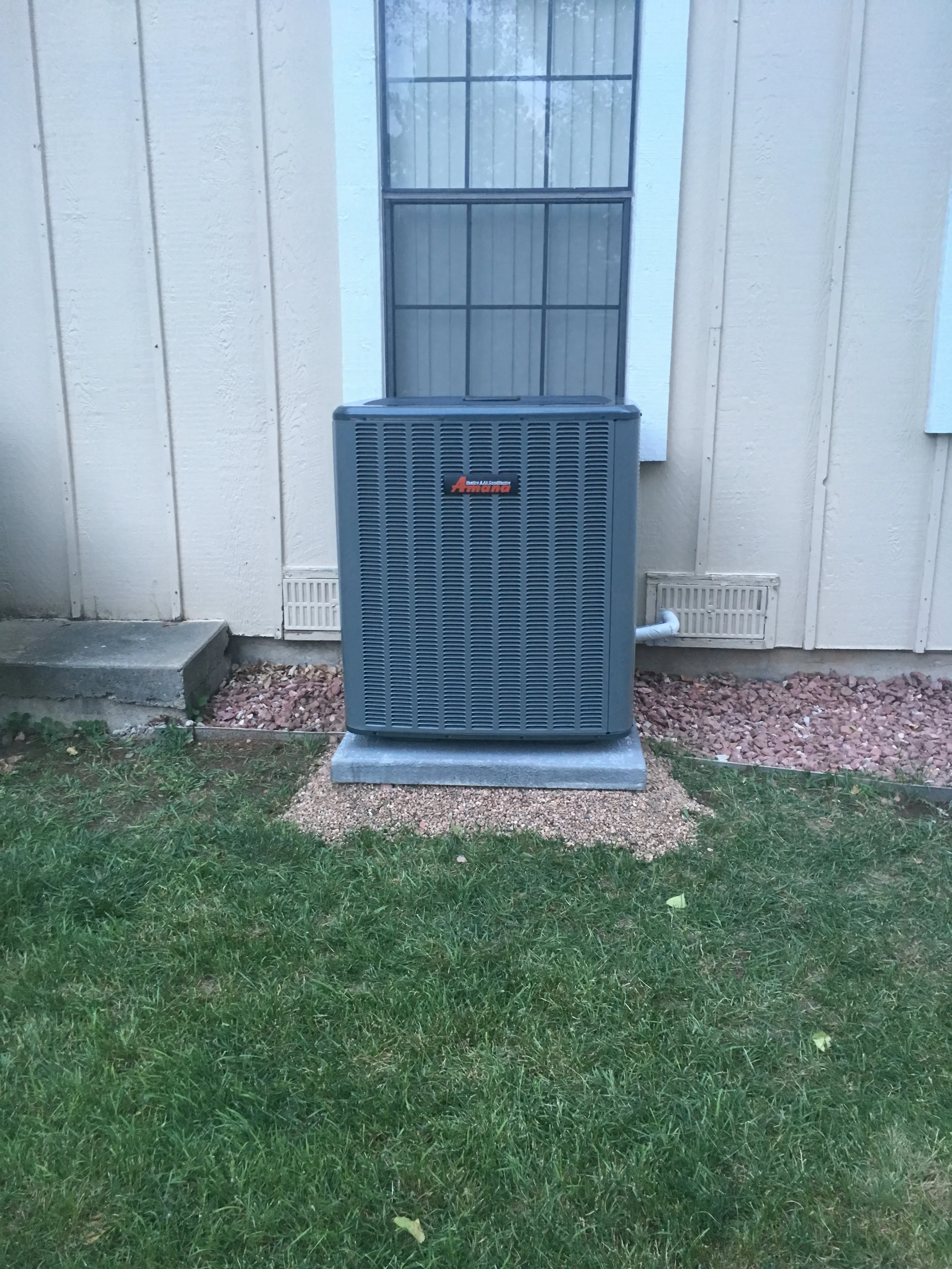 Outdoor view of an air conditioning unit installed against the exterior wall of a house, with a window above and a small gravel area underneath.