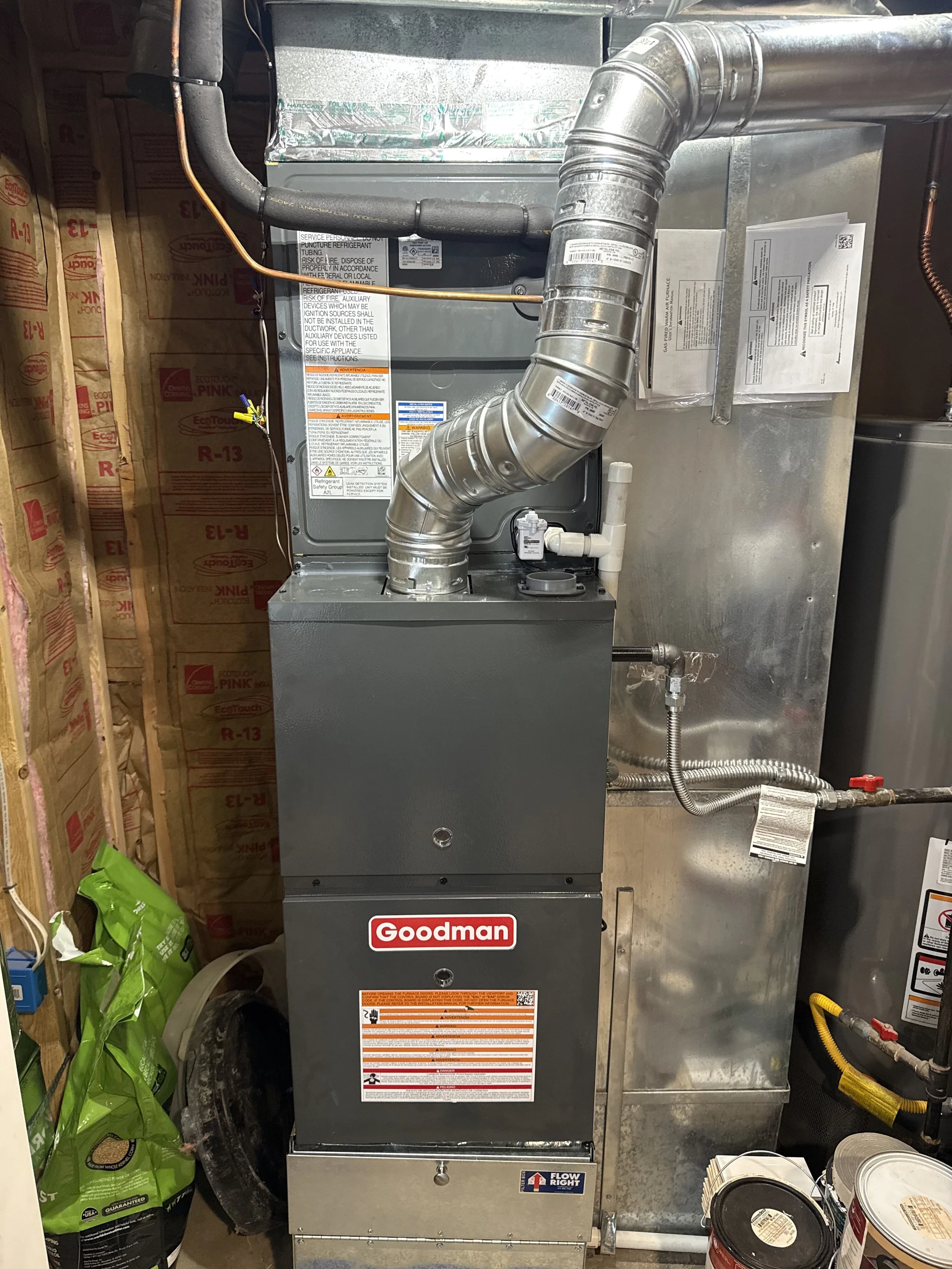 A Goodman HVAC furnace and ductwork in a utility room with insulation on the walls, pipes, and other mechanical components.