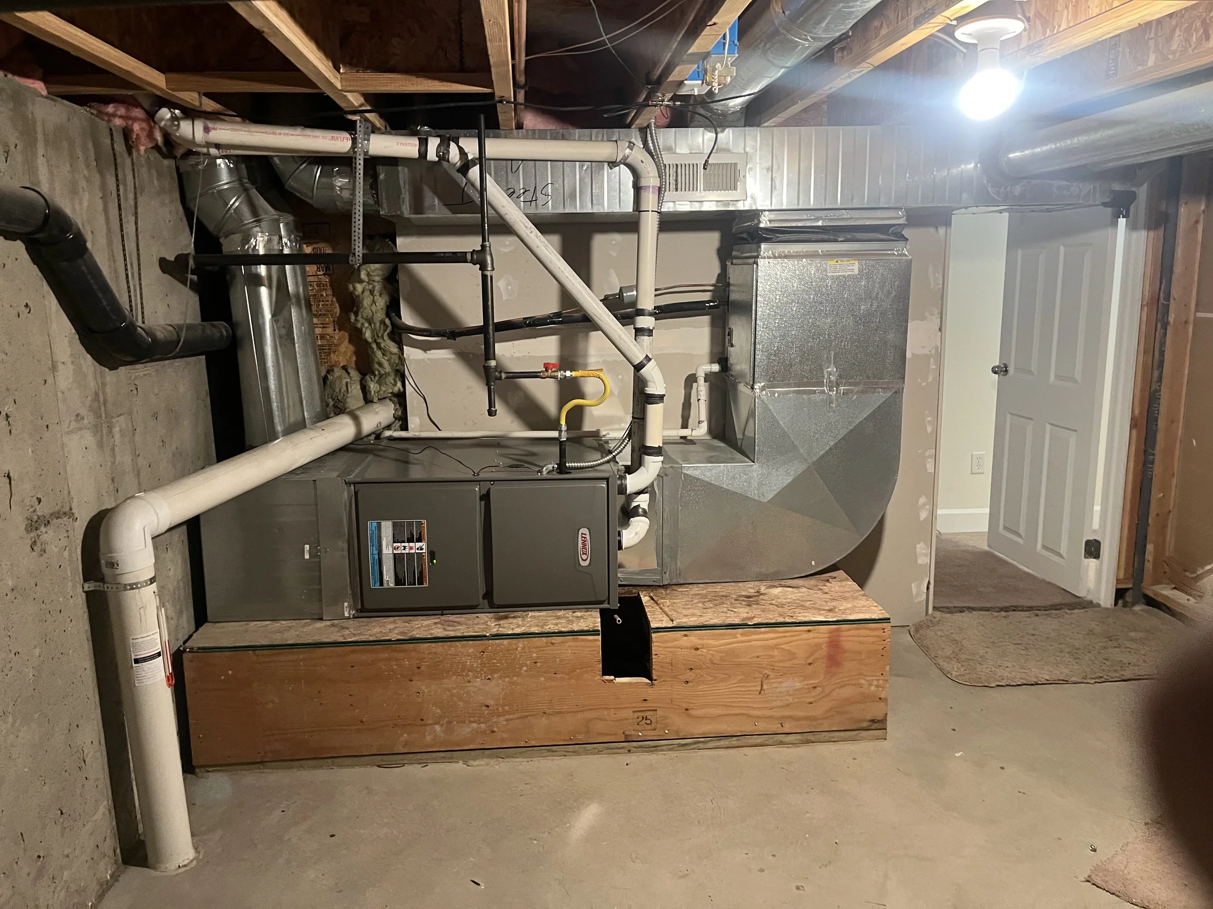 A view of a furnace and ductwork system in an unfinished basement. The furnace is metallic gray with piping connected to it and a wooden platform underneath. The basement floor is concrete, and there is a white door with a doormat in the background. 