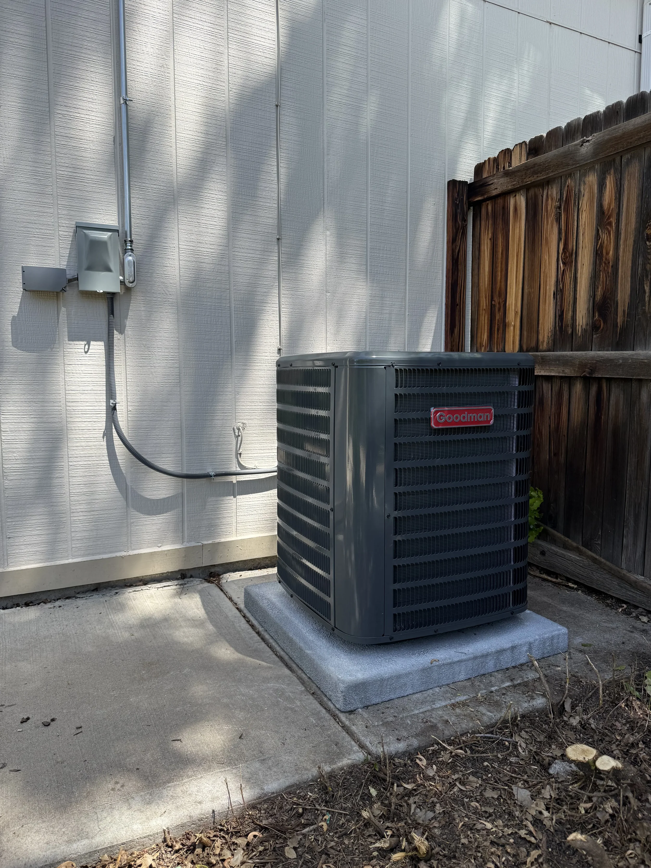 An outdoor air conditioning unit by Goodman, situated on a concrete pad next to a wooden fence and a house wall with utility connections.