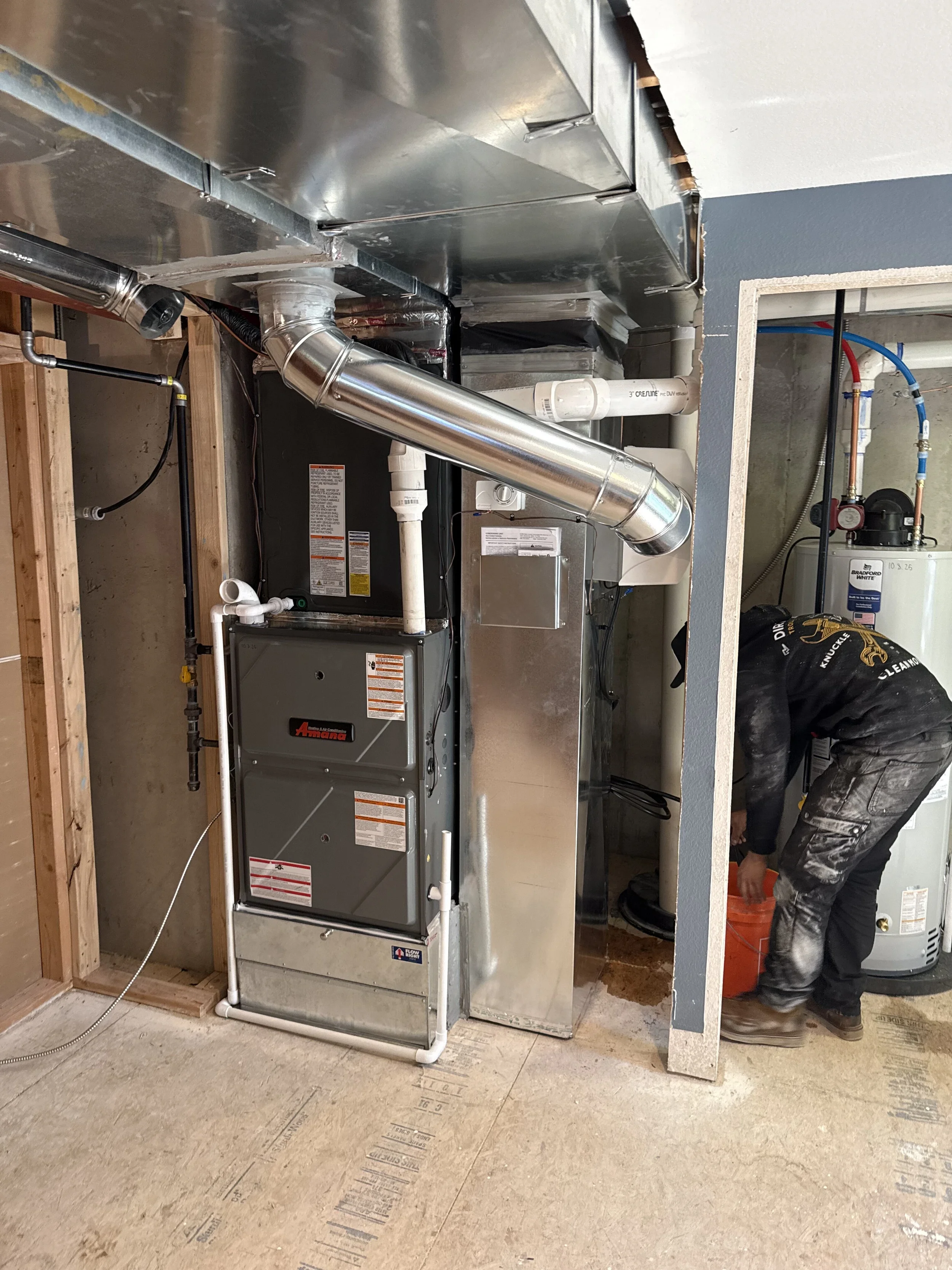A worker installing or repairing HVAC equipment in a utility room with pipes, ductwork, and a water heater.