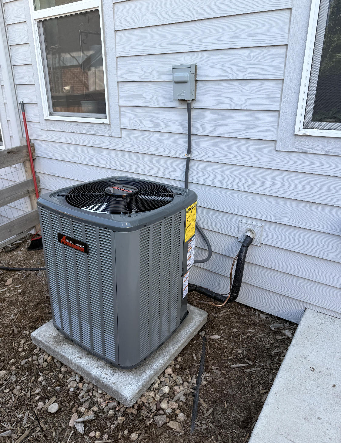 Outdoor air conditioning unit installed on a concrete slab next to the house wall, with electrical connections and a nearby window.