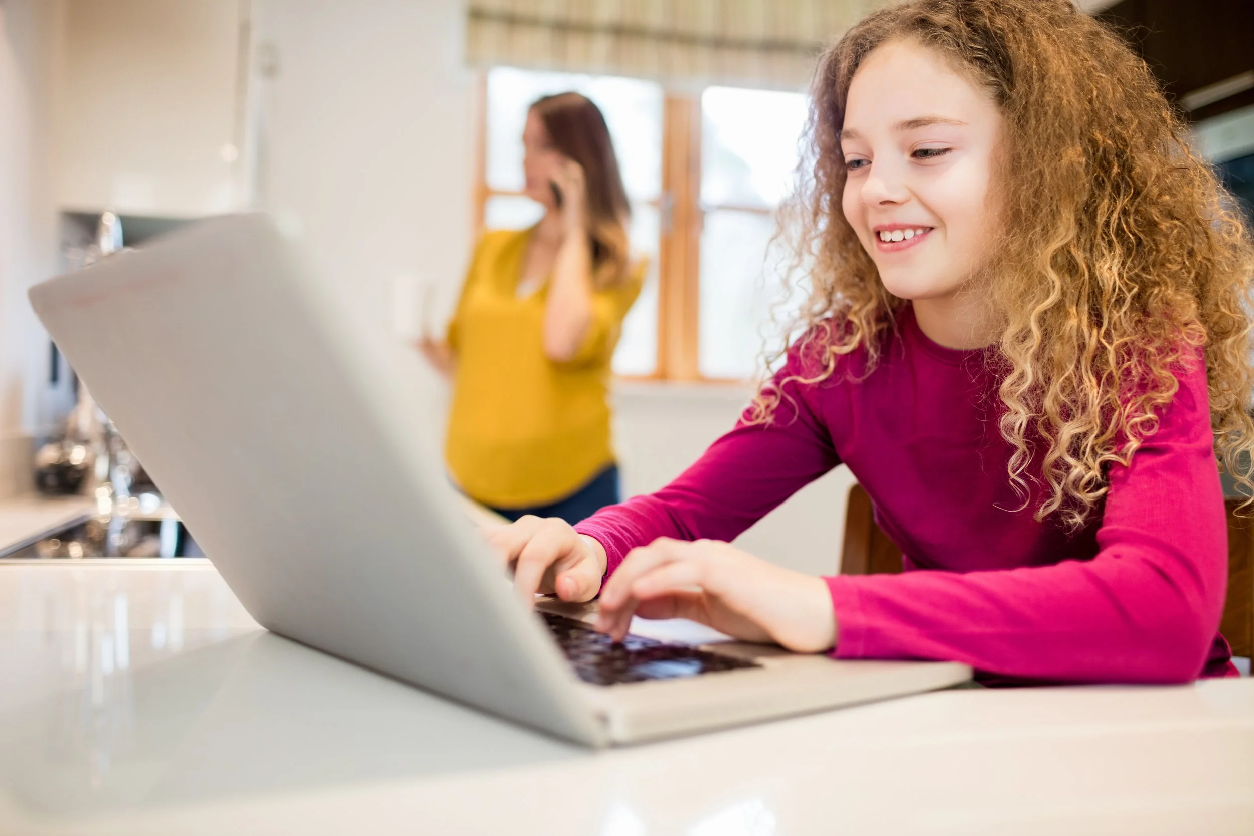 Young girl with curly hair smiling while using a laptop at a kitchen table, woman in yellow shirt talking on the phone in background.