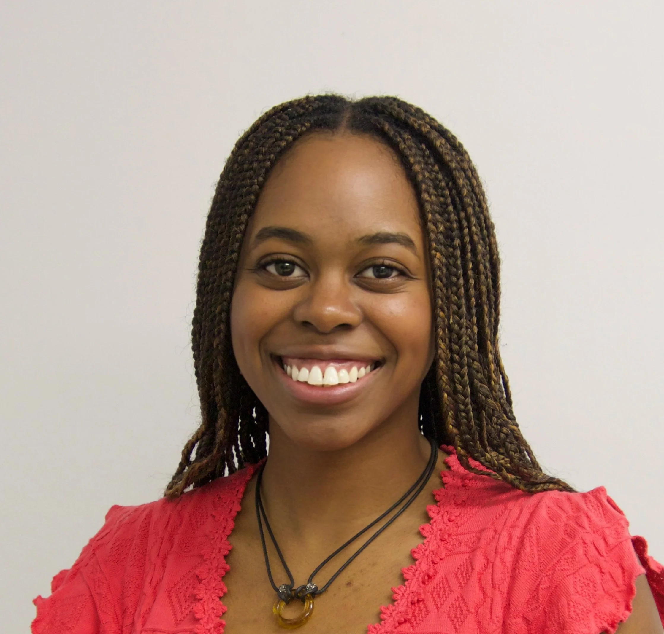 A smiling woman with braided hair wearing a red blouse and a black necklace with a gold ring pendant.