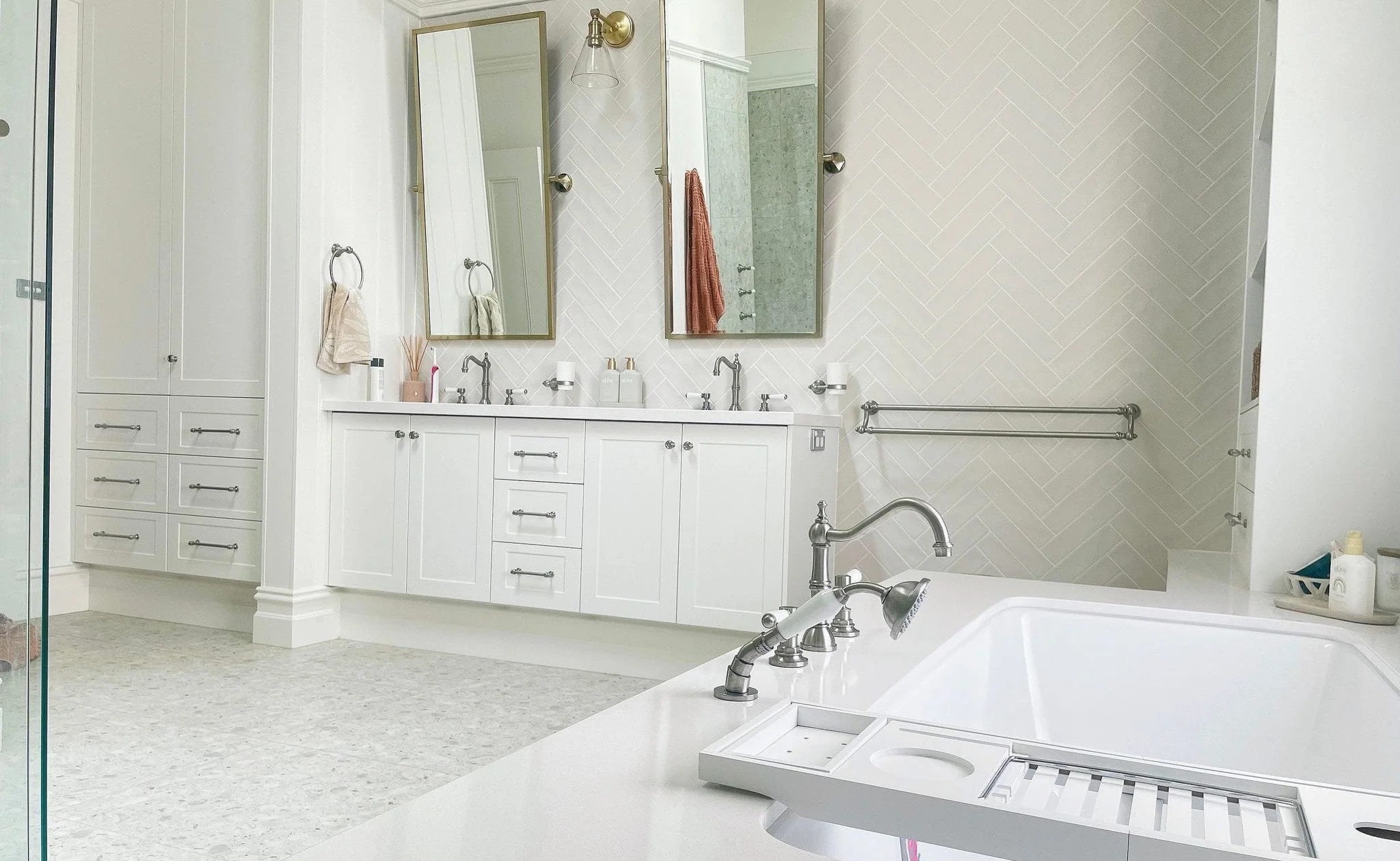 Modern white bathroom with double vanity, two large mirrors, and a bathtub. Towels hang on the wall, and various toiletries are on the counter. A shower stall is visible on the left.