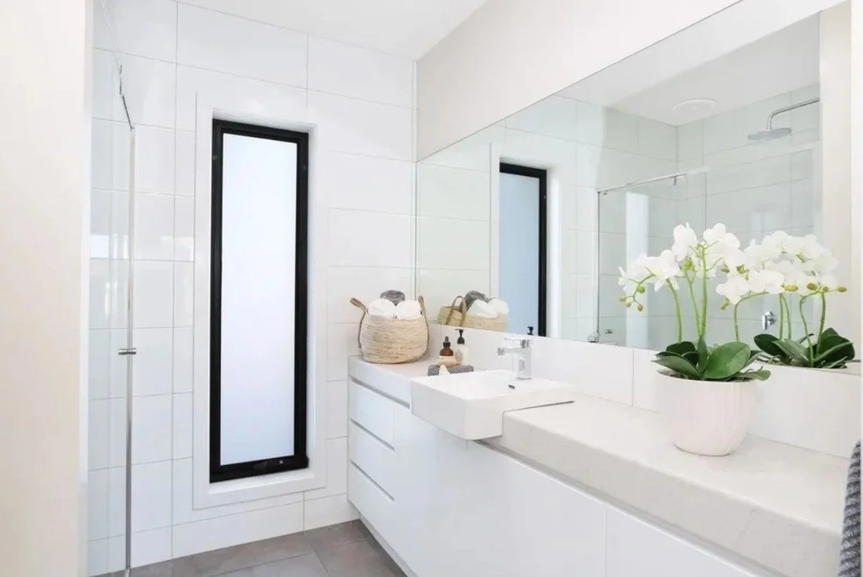 A modern bathroom with white tiles, a large mirror, black-framed window, white vanity with a vessel sink, a potted orchid, and wicker baskets on the countertop.