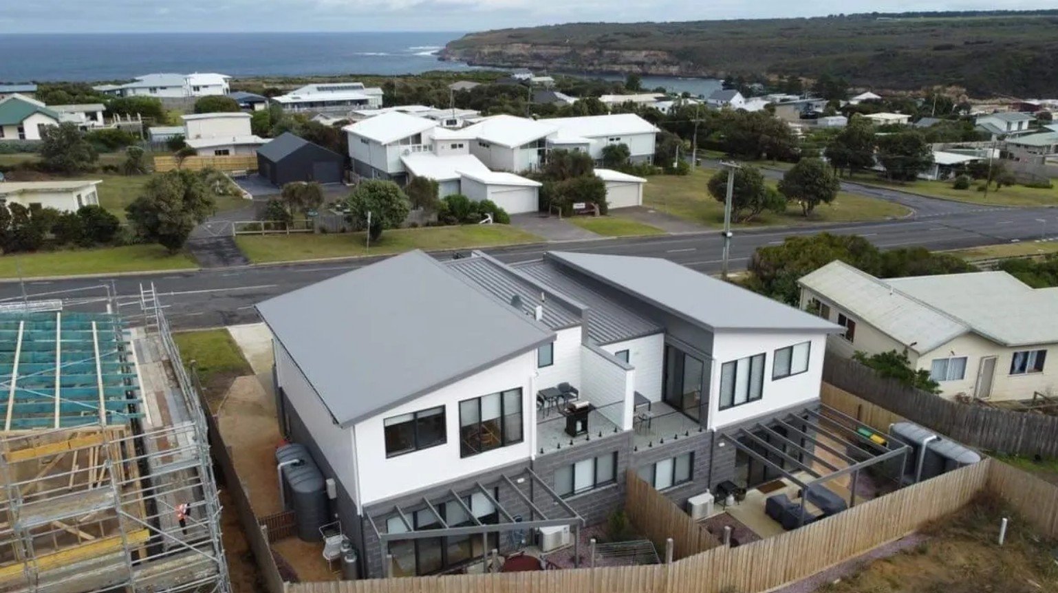 Aerial view of modern houses near the coastline with grassy hills and ocean in the background.