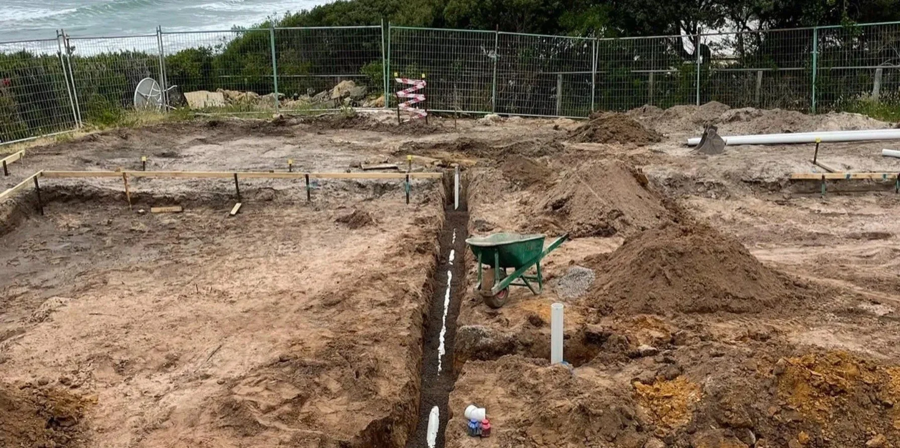 Construction site showing trenches, dirt piles, PVC pipes, and a wheelbarrow on an overcast day.