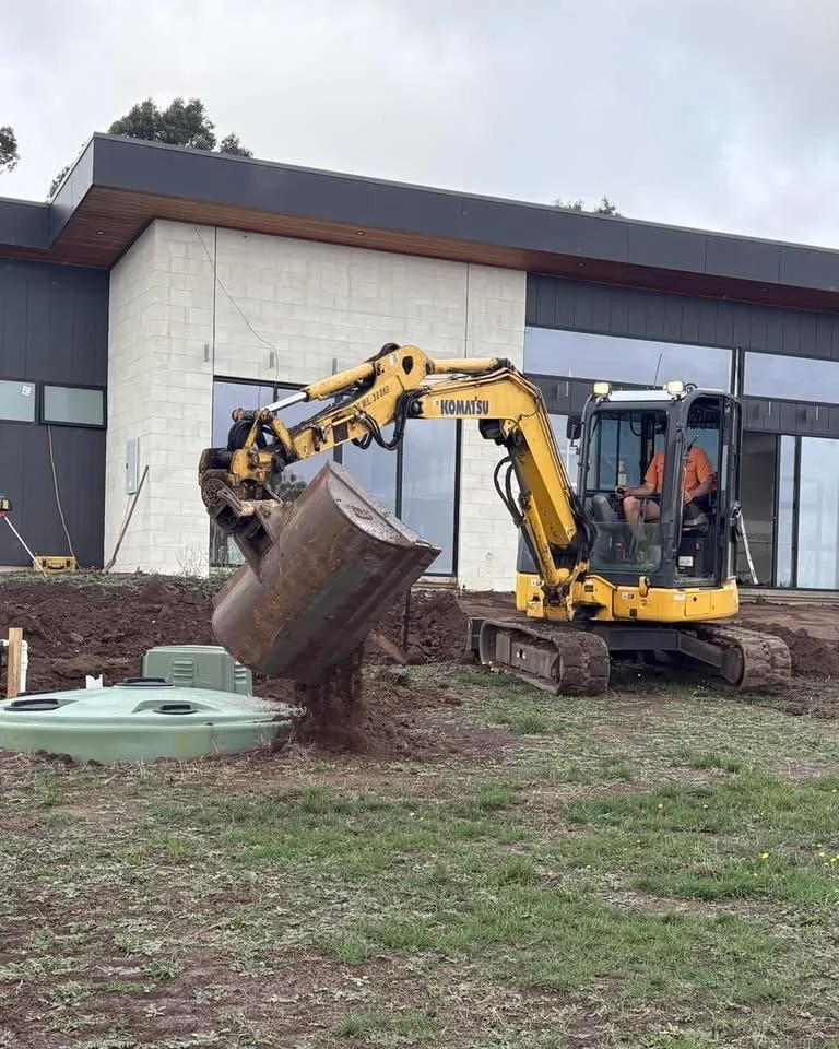 A yellow Komatsu mini excavator digging in front of a modern building with gray and white exterior walls.