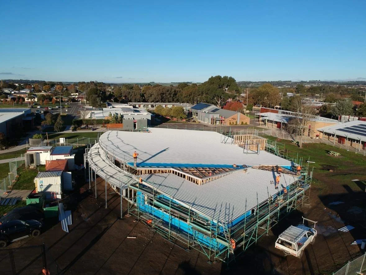Construction site of a facility with a curved roof nearing completion, workers installing roofing materials, surrounded by trees and small buildings.