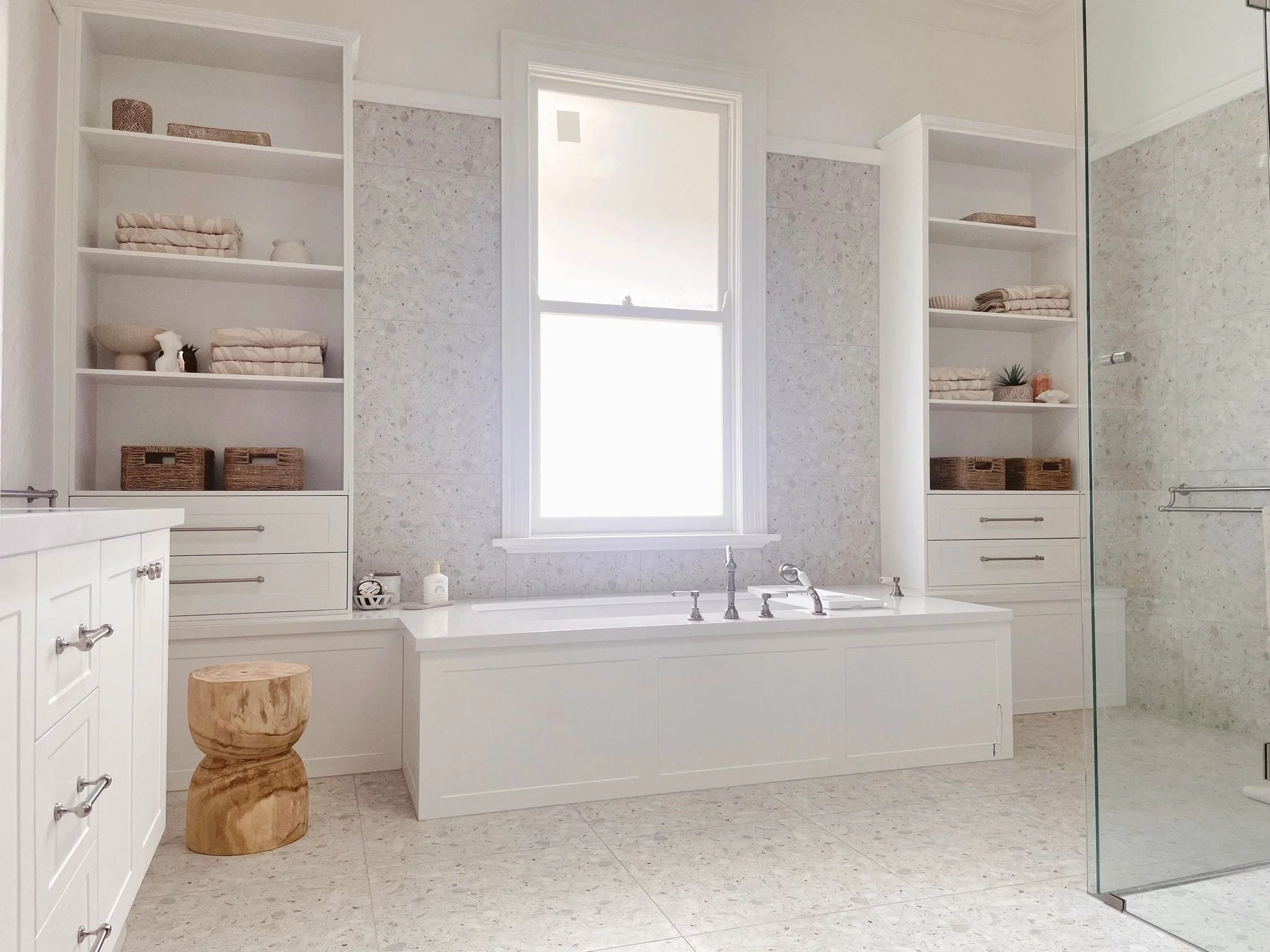 Bright bathroom featuring a built-in bathtub beneath a large window, with open shelves on either side filled with towels, decor, and baskets. A wooden stool is positioned near the vanity, and a glass shower enclosure extends to the right.