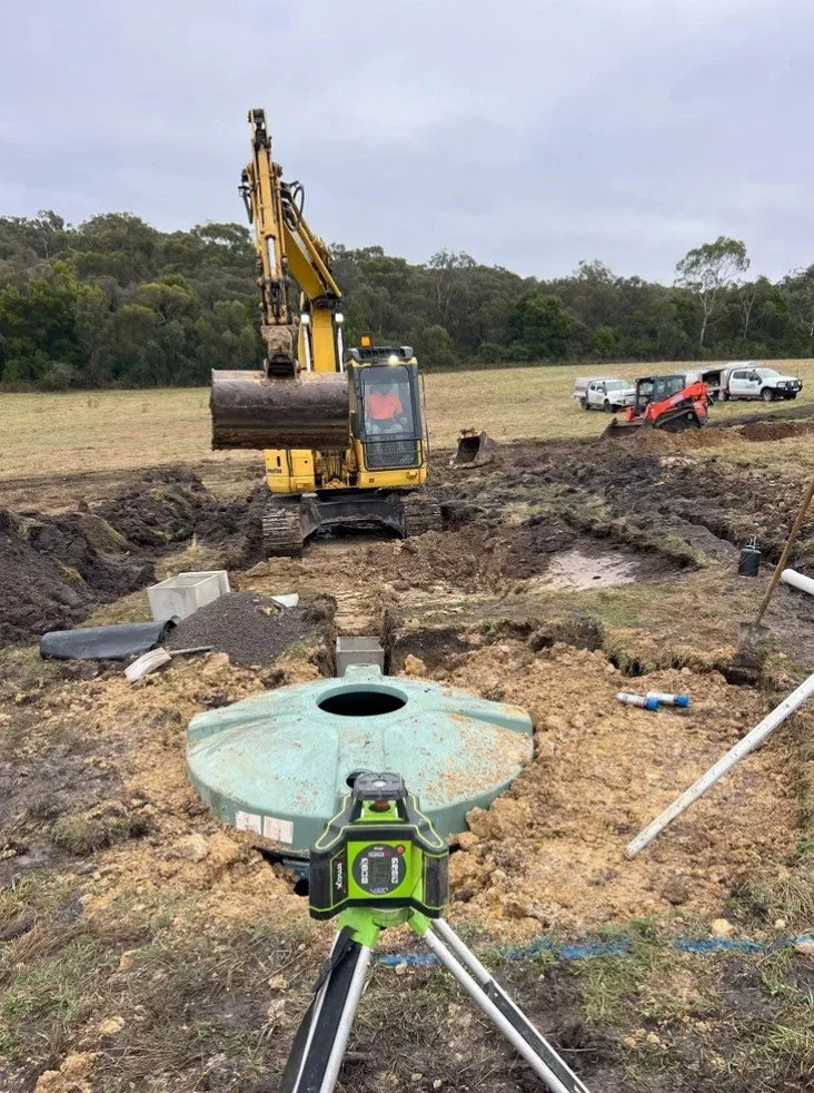 Construction site with an excavator digging a trench for underground utility installation, including a green septic tank cover, surveying equipment, and parked vehicles in the background.