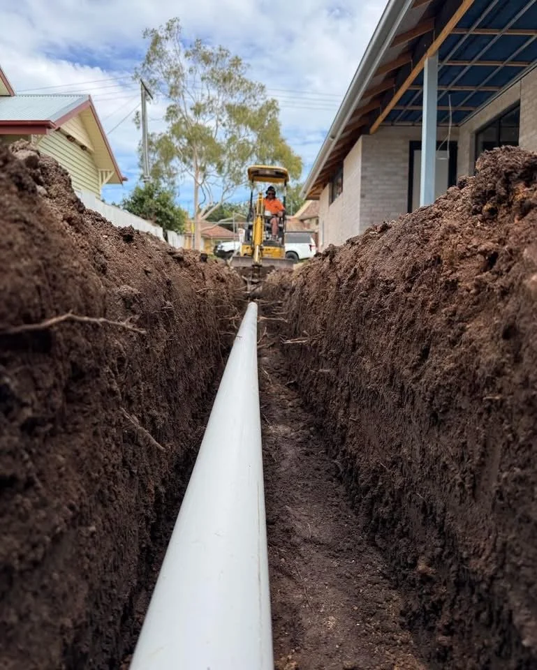 A construction worker operating a small excavator digs a trench in a residential area for installing a large pipe.