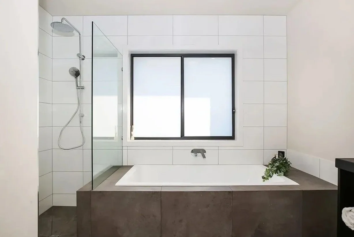 Modern bathroom with a bathtub beneath a window, a walk-in shower with glass partition, white tiled walls, and a black vanity on the right side. A green plant rests on the edge of the bathtub.