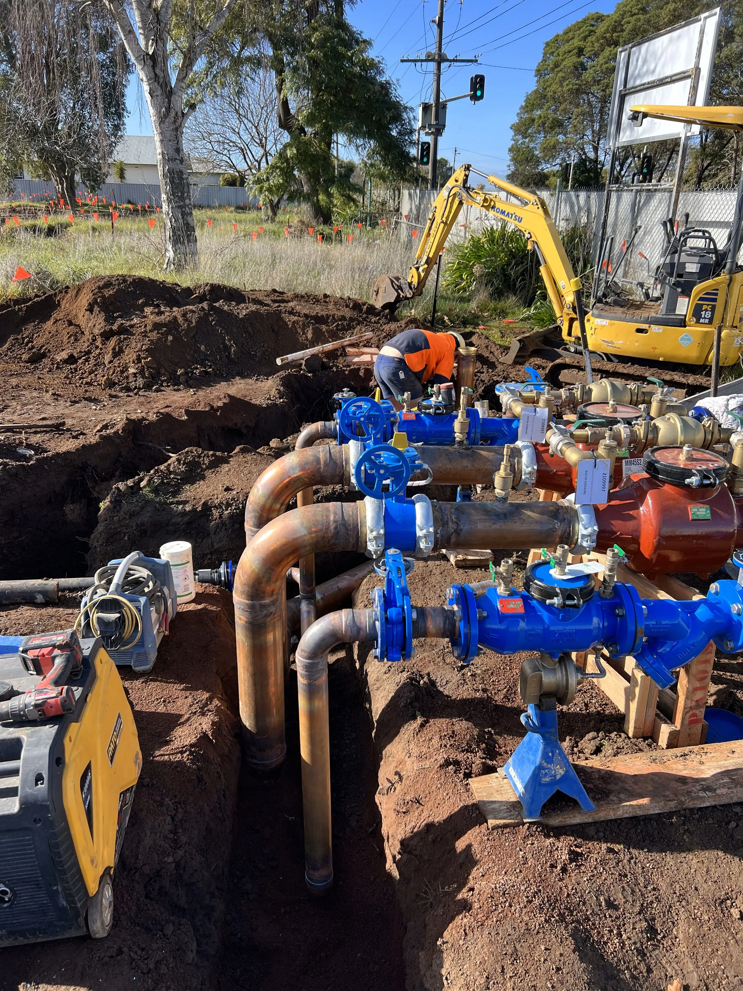 Construction worker in orange shirt working on industrial pipes with blue valves, surrounded by dirt and construction equipment, under a clear sunny sky.