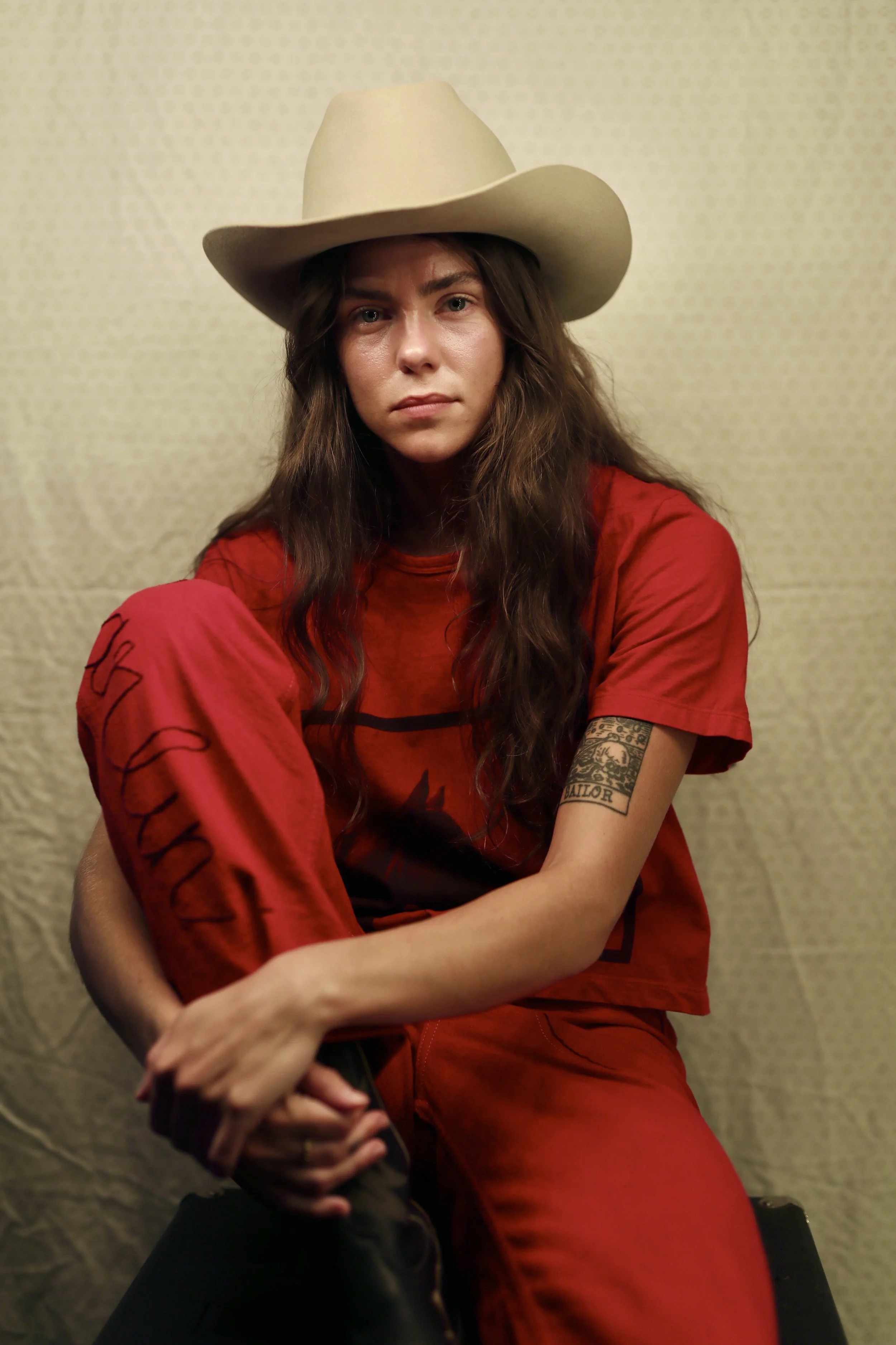 Young woman with long wavy hair wearing a tan cowboy hat, red t-shirt, and red pants, sitting against a beige textured background.