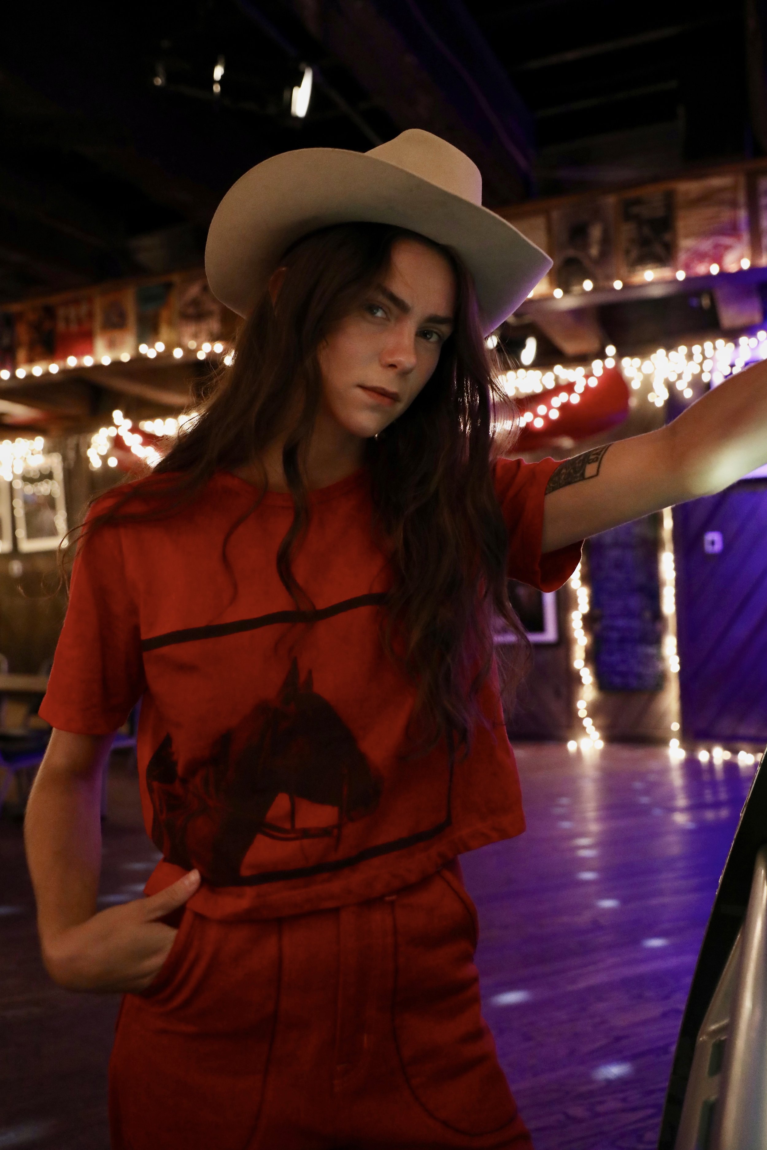 A young woman with long dark hair wearing a beige hat and red graphic t-shirt standing in a dimly lit room with string lights, wooden walls, and arcade machine.