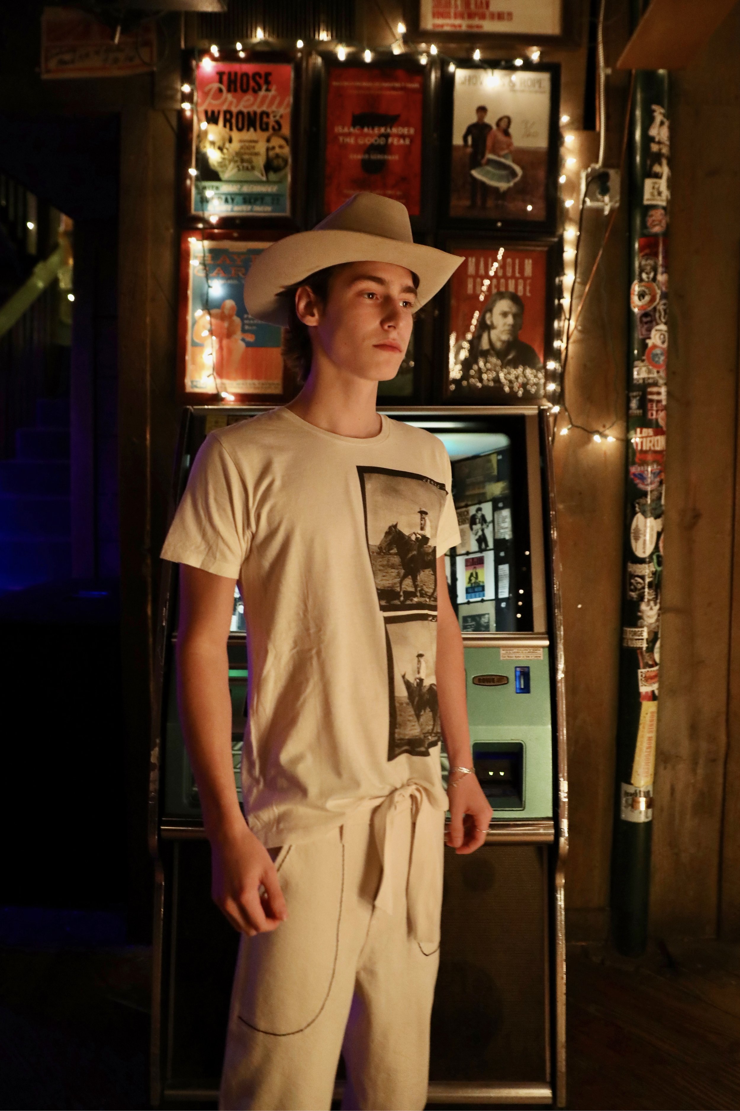 A young person standing in front of a vintage soda vending machine in a dimly lit room decorated with posters and string lights, wearing a beige T-shirt with a graphic print and matching beige sweatpants, along with a wide brim hat.