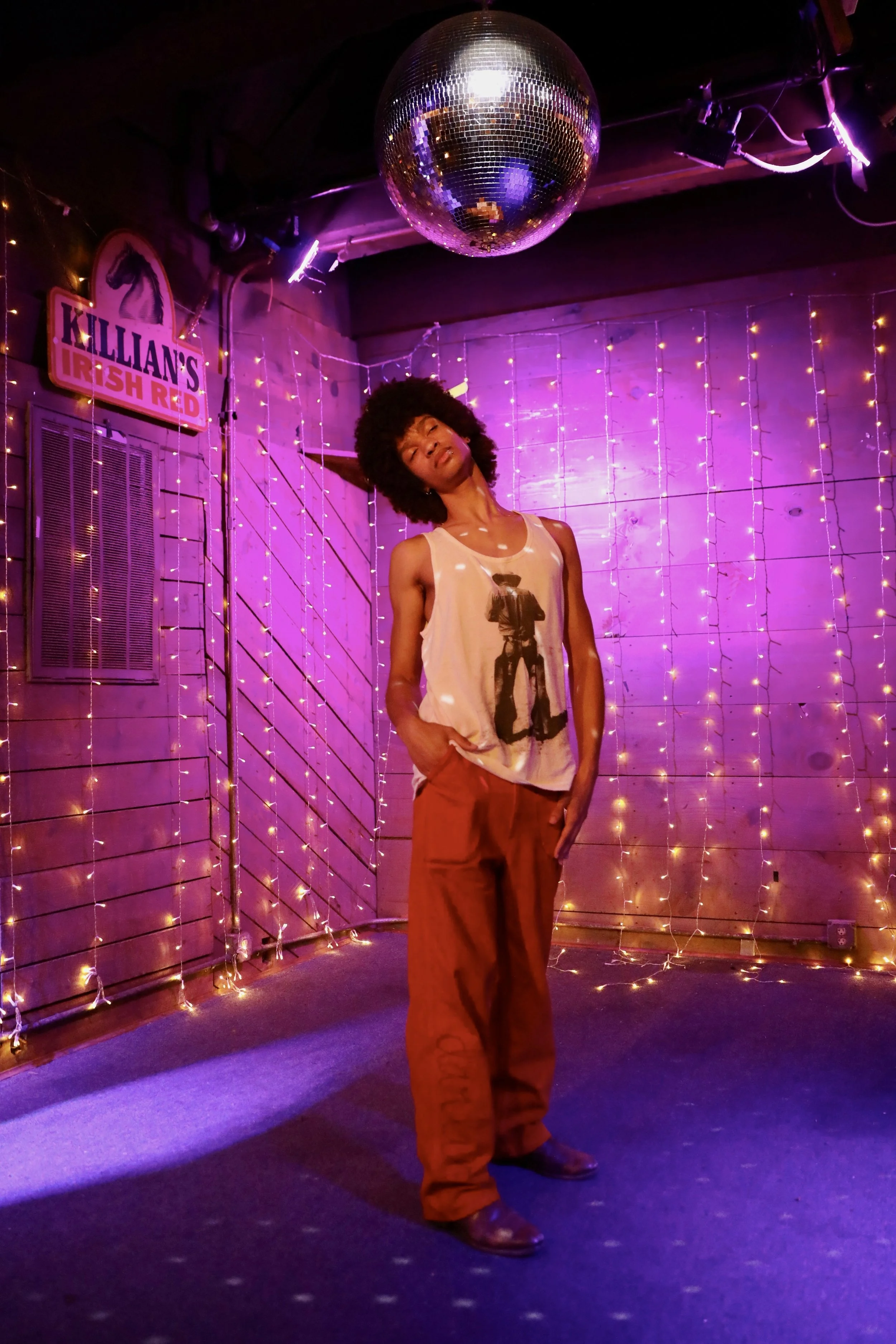 A person with curly hair standing in a colorful, neon-lit club with purple and pink string lights, a disco ball hanging from the ceiling, and a sign that reads 'Killian's Irish Red' on the wall.