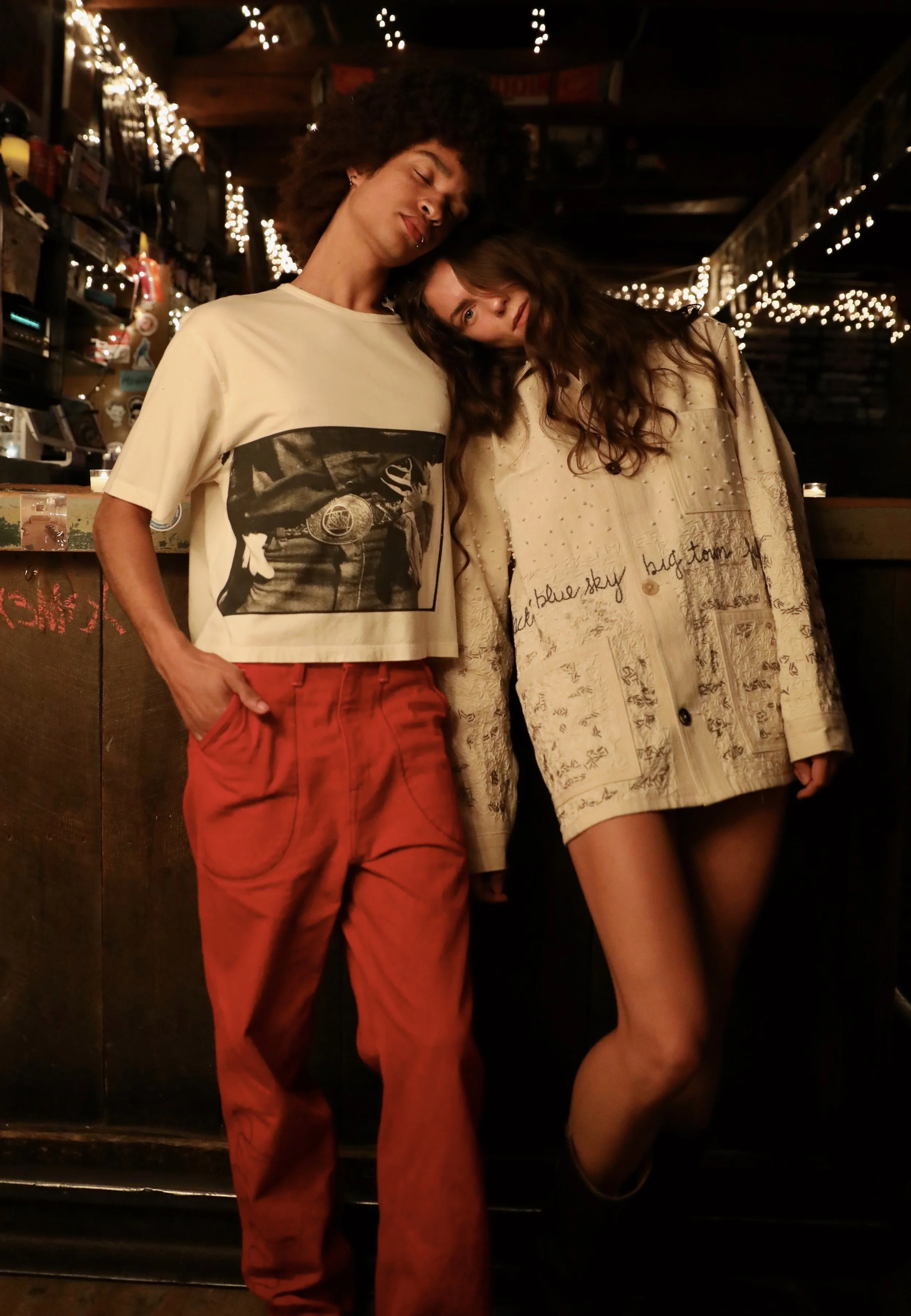 Two young women standing close together, leaning their heads against each other, in a cozy indoor setting with string lights. One wears a cream-colored jacket with handwritten words and designs, the other wears a cream t-shirt with a printed image and red pants.