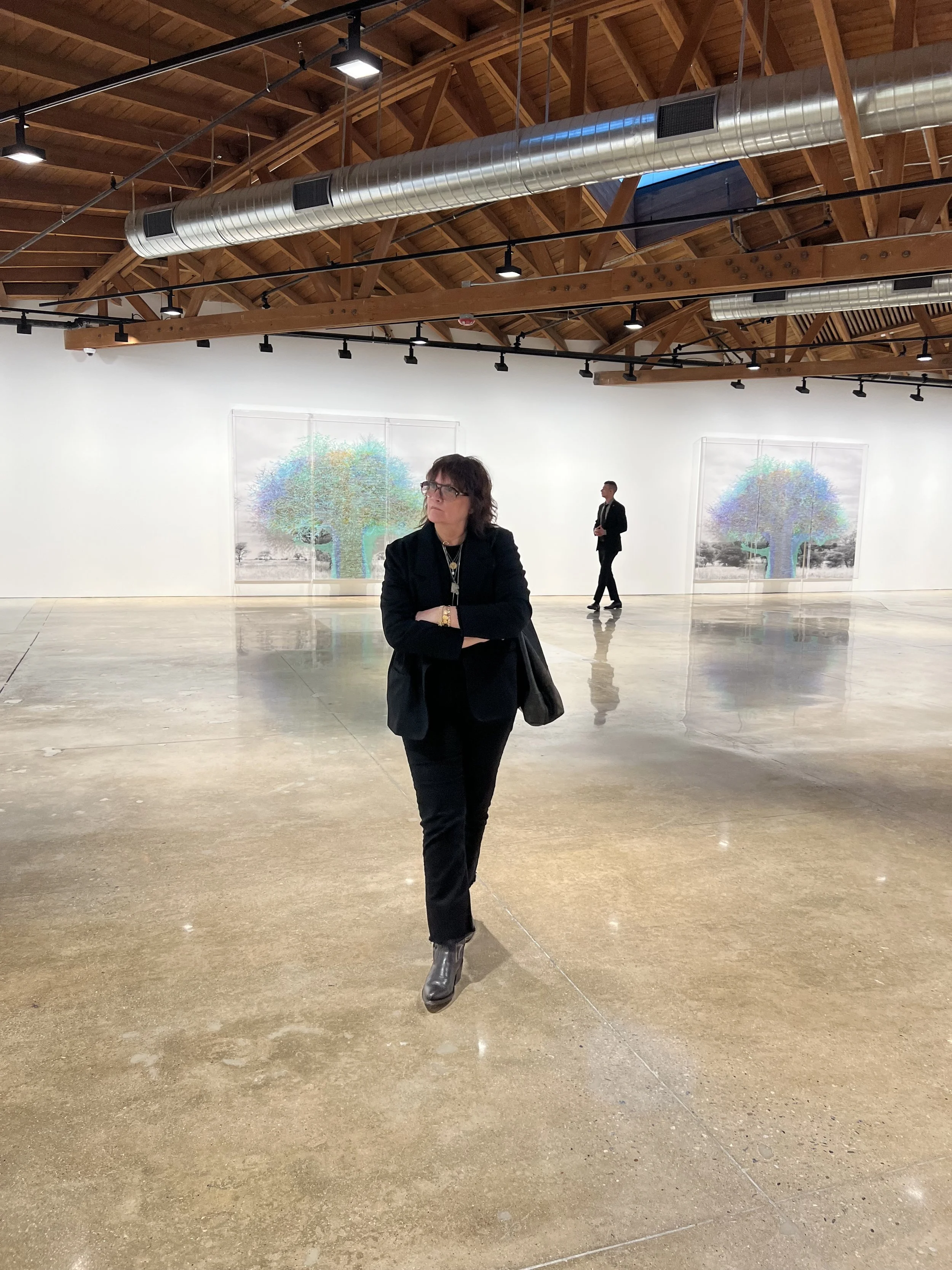 A woman standing with arms crossed in an art gallery with colorful trees displayed in the background and a man walking in the distance.