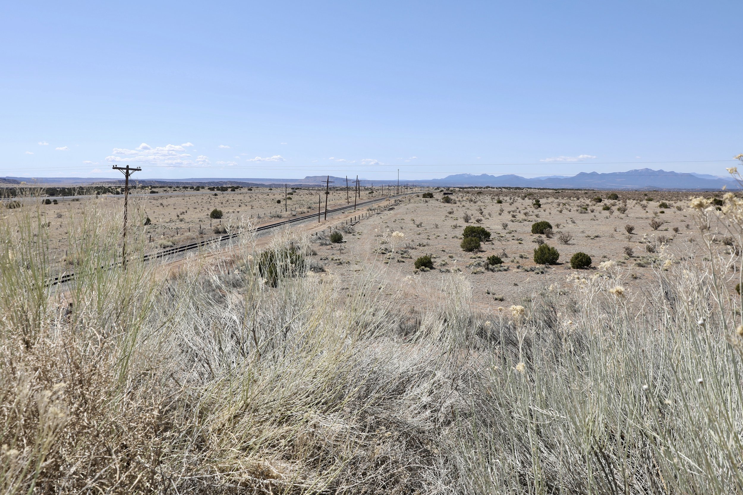 A vast desert landscape with sparse vegetation, railroad tracks running through the middle, electrical poles alongside the tracks, and distant mountains under a blue sky with few clouds.