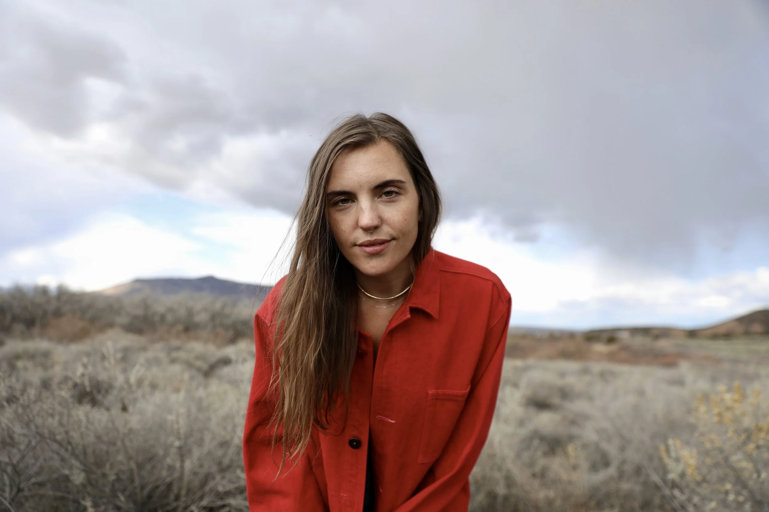 A young woman with long brown hair and light skin, wearing a red jacket, stands outdoors in a desert landscape with mountains in the distance under cloudy skies.