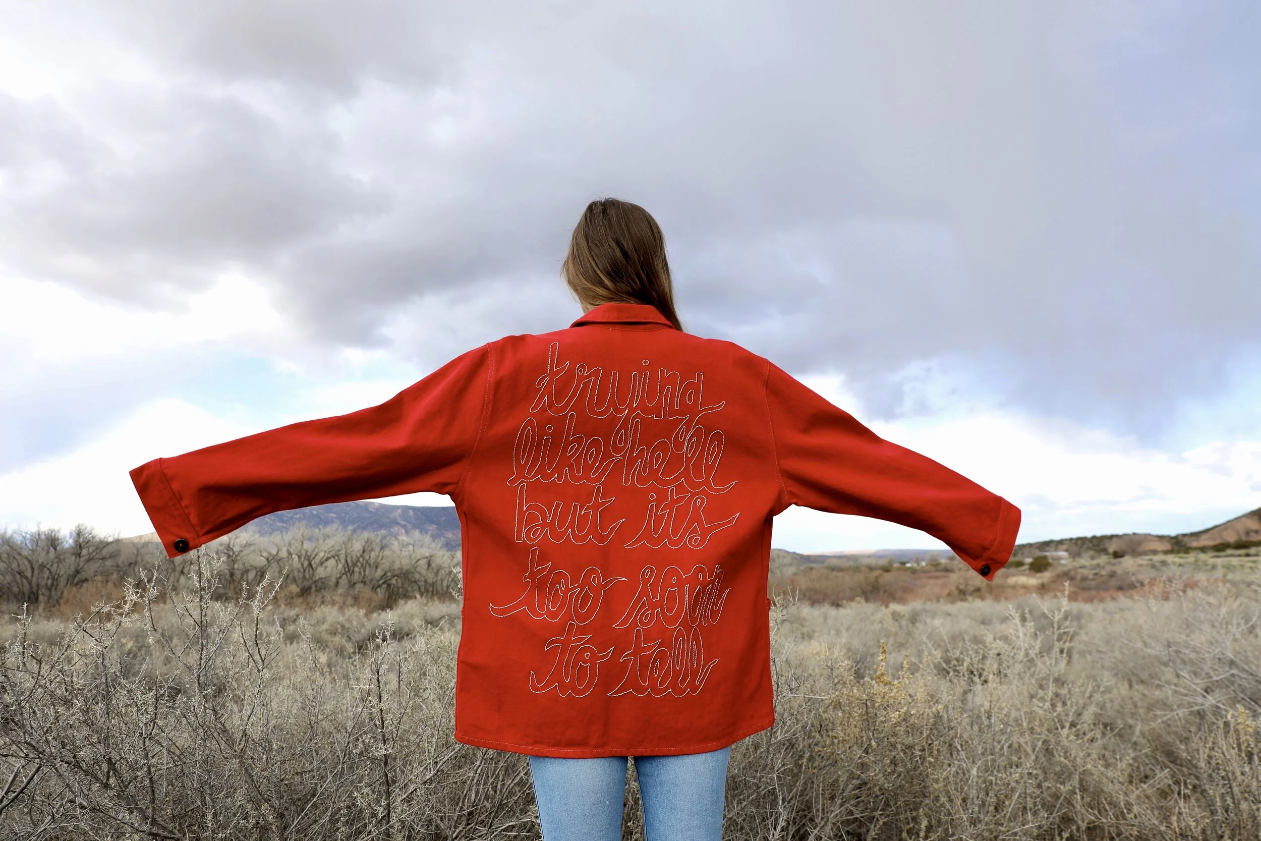 Back view of a woman wearing a red jacket with embroidered text, standing in an open natural landscape with cloudy sky.