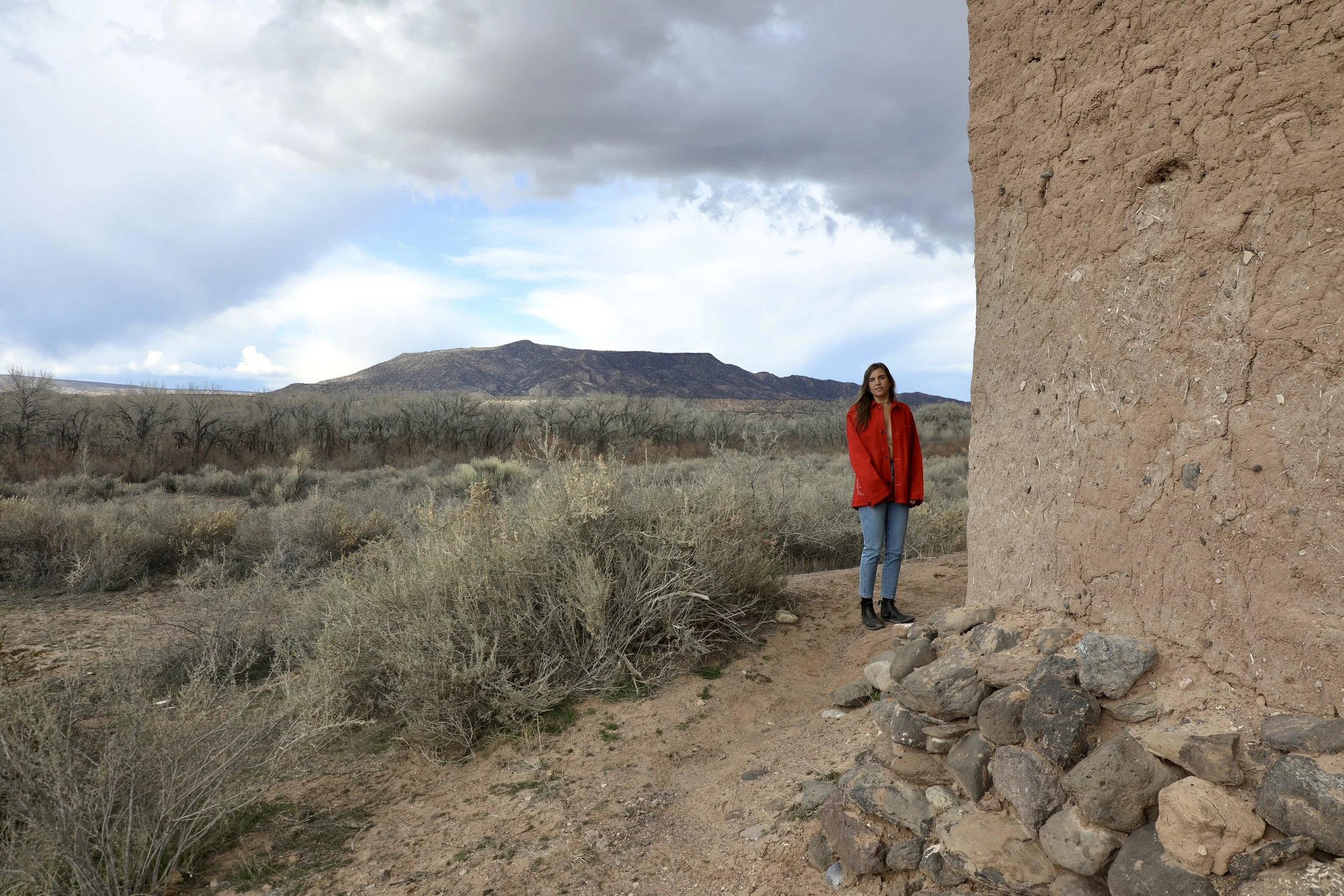 A woman standing beside a large adobe wall in a desert landscape with mountains in the background. She is wearing a red coat, jeans, and black boots. The sky is partly cloudy.