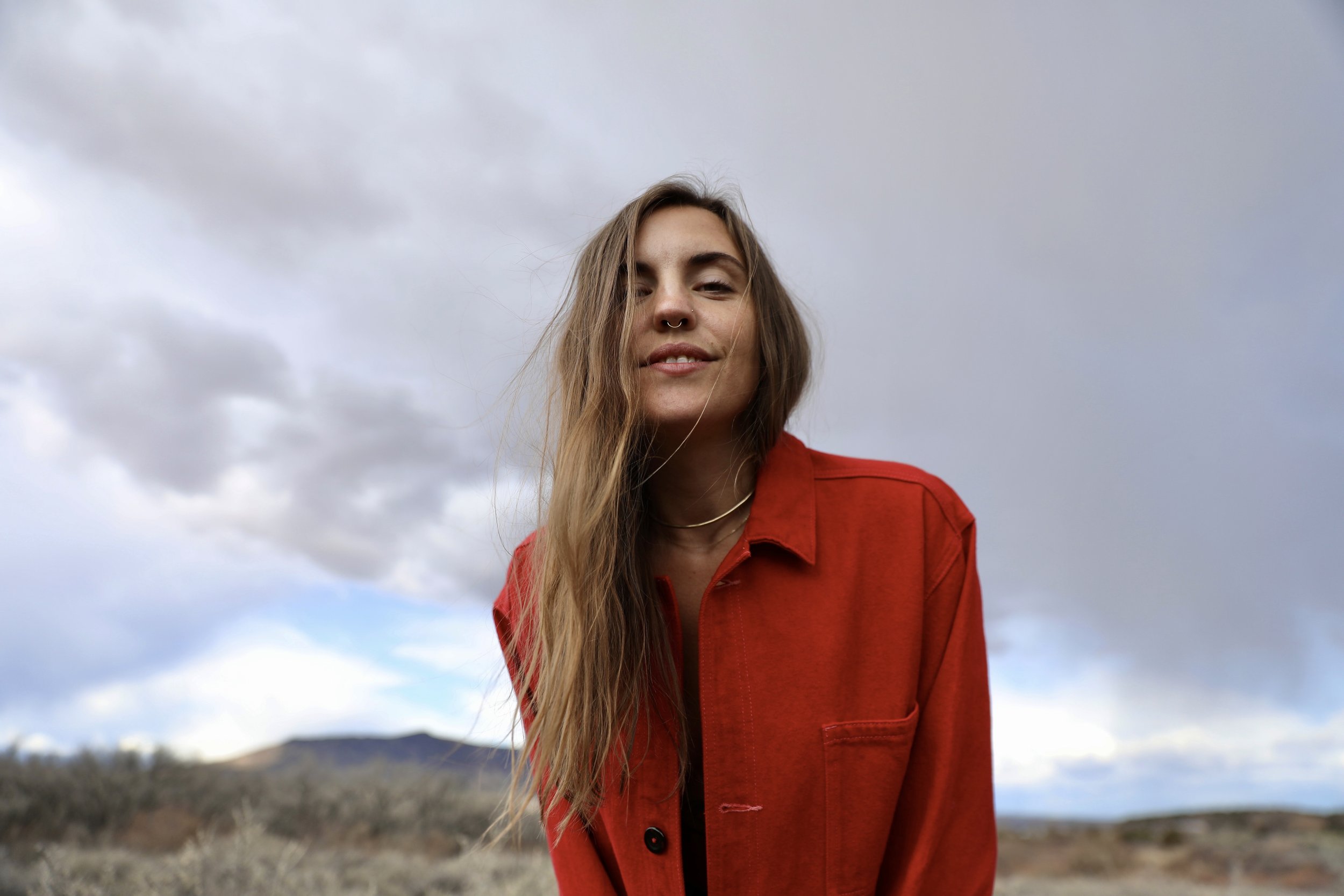 A young woman with long hair and a septum piercing wearing a red jacket outdoors under cloudy skies in a desert landscape.