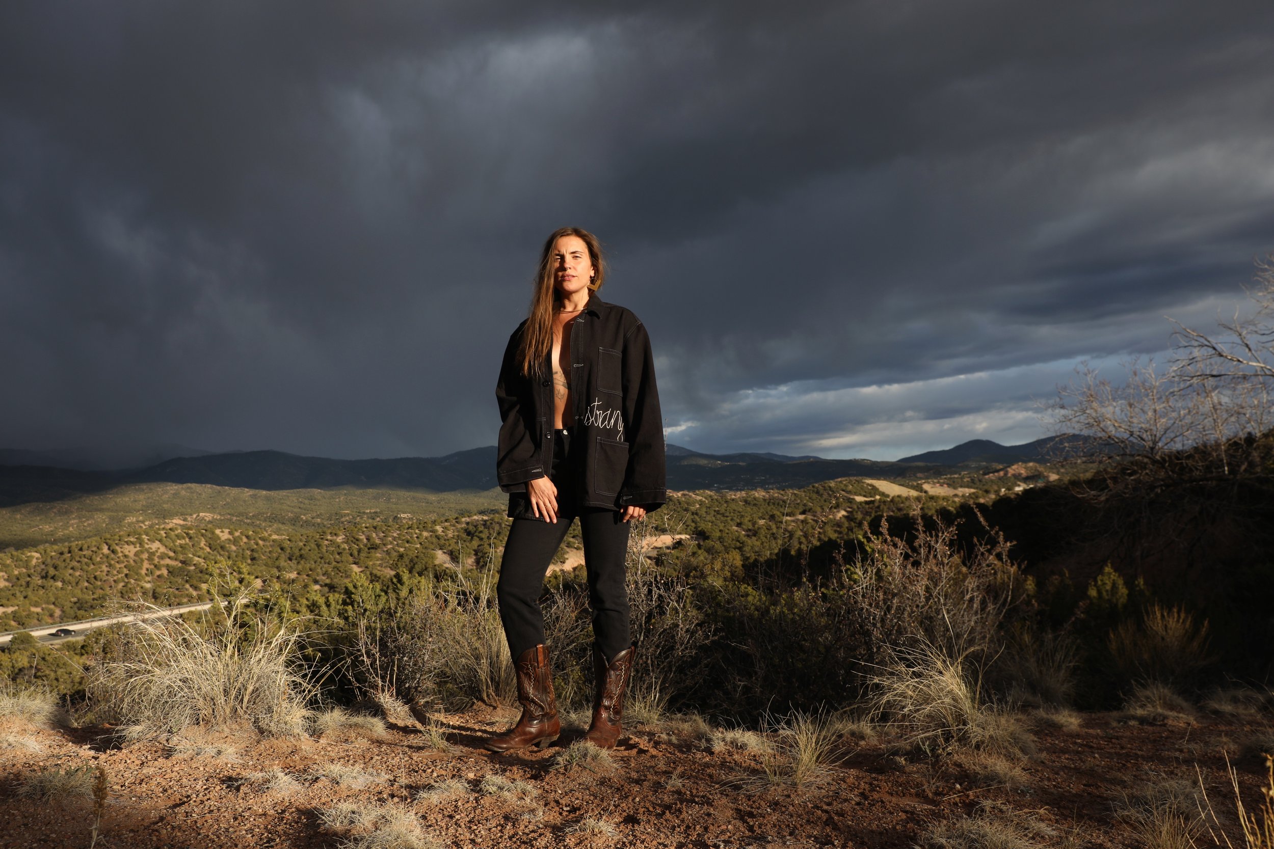 A woman standing outdoors on a hilltop with dark stormy clouds in the sky behind her, wearing a black jacket, black pants, and brown cowboy boots.