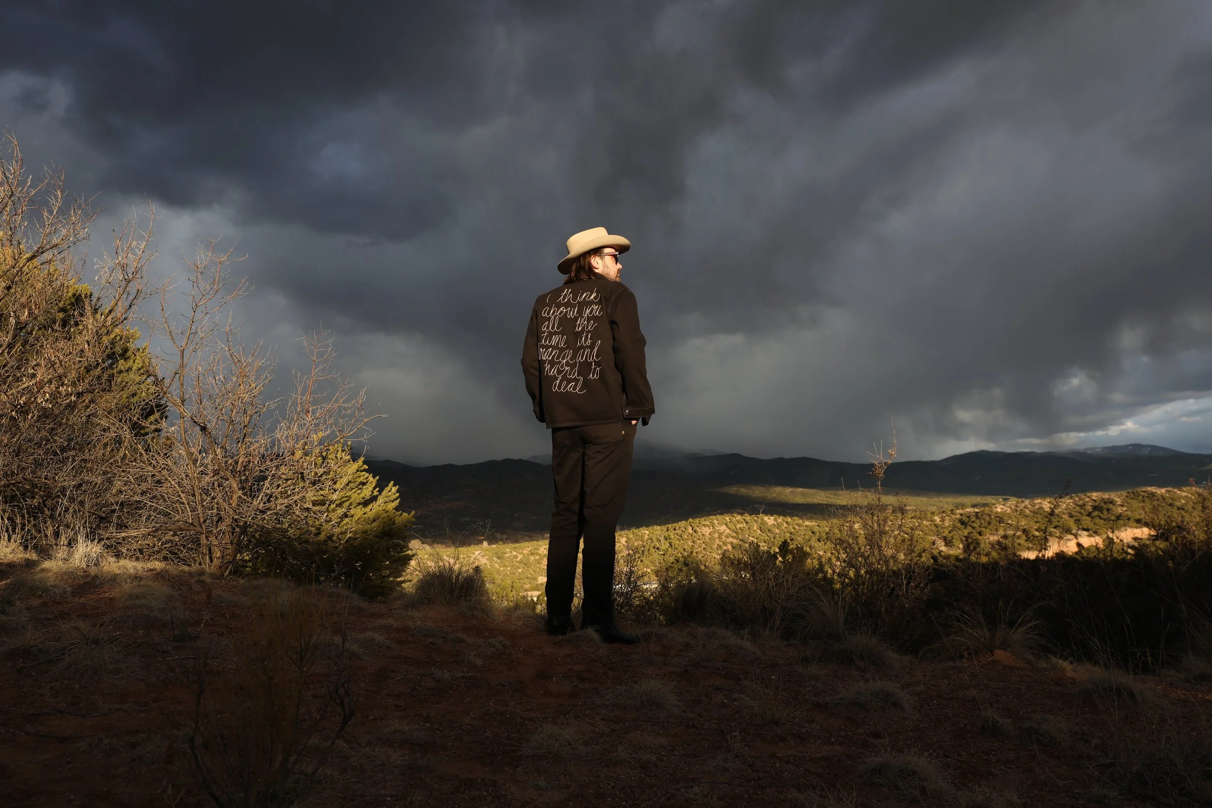A person wearing a beige hat and a dark jacket with writing on the back, standing on a hill under dark storm clouds and looking at the distant mountains.