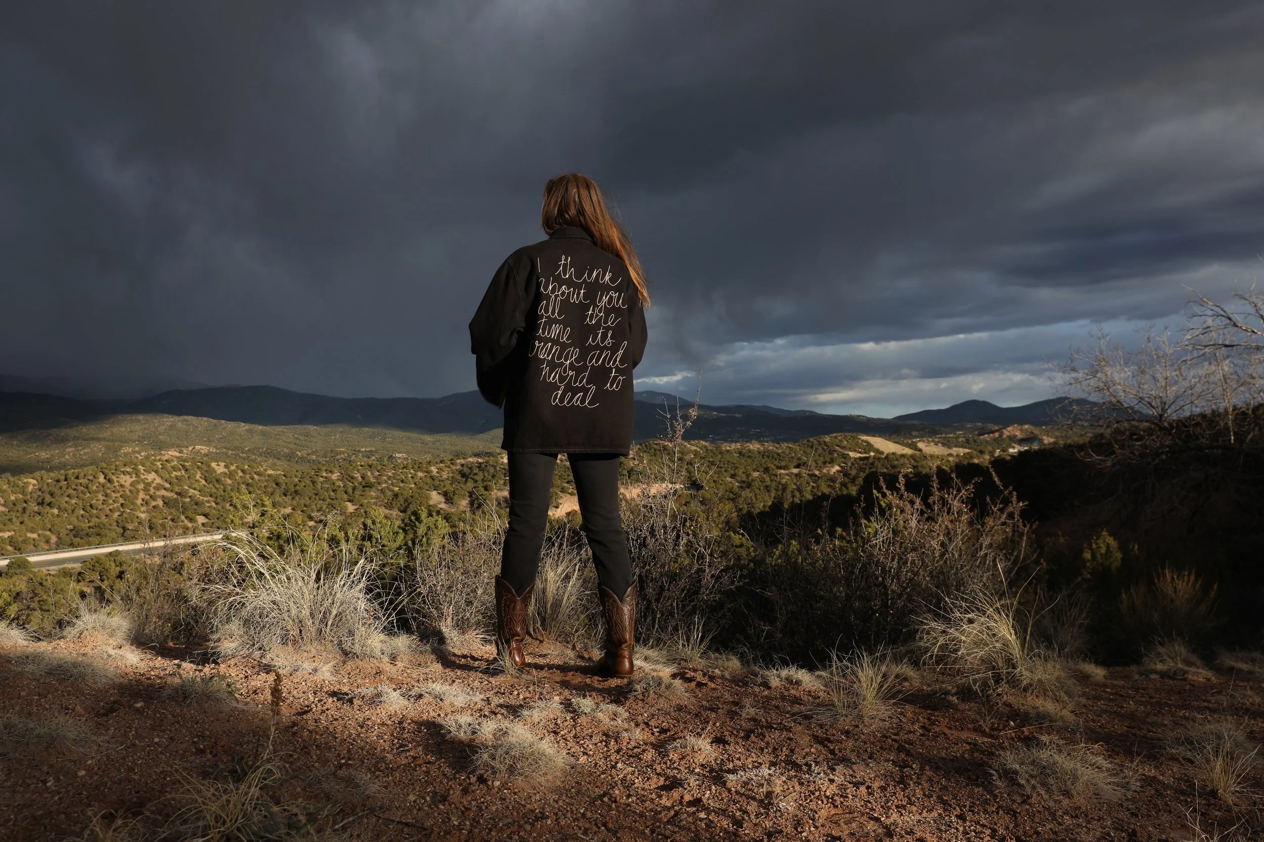 A woman standing on a dirt hill overlooking a vast landscape of rolling hills and mountains under a dark, stormy sky, wearing a black jacket with writing on the back and cowboy boots.