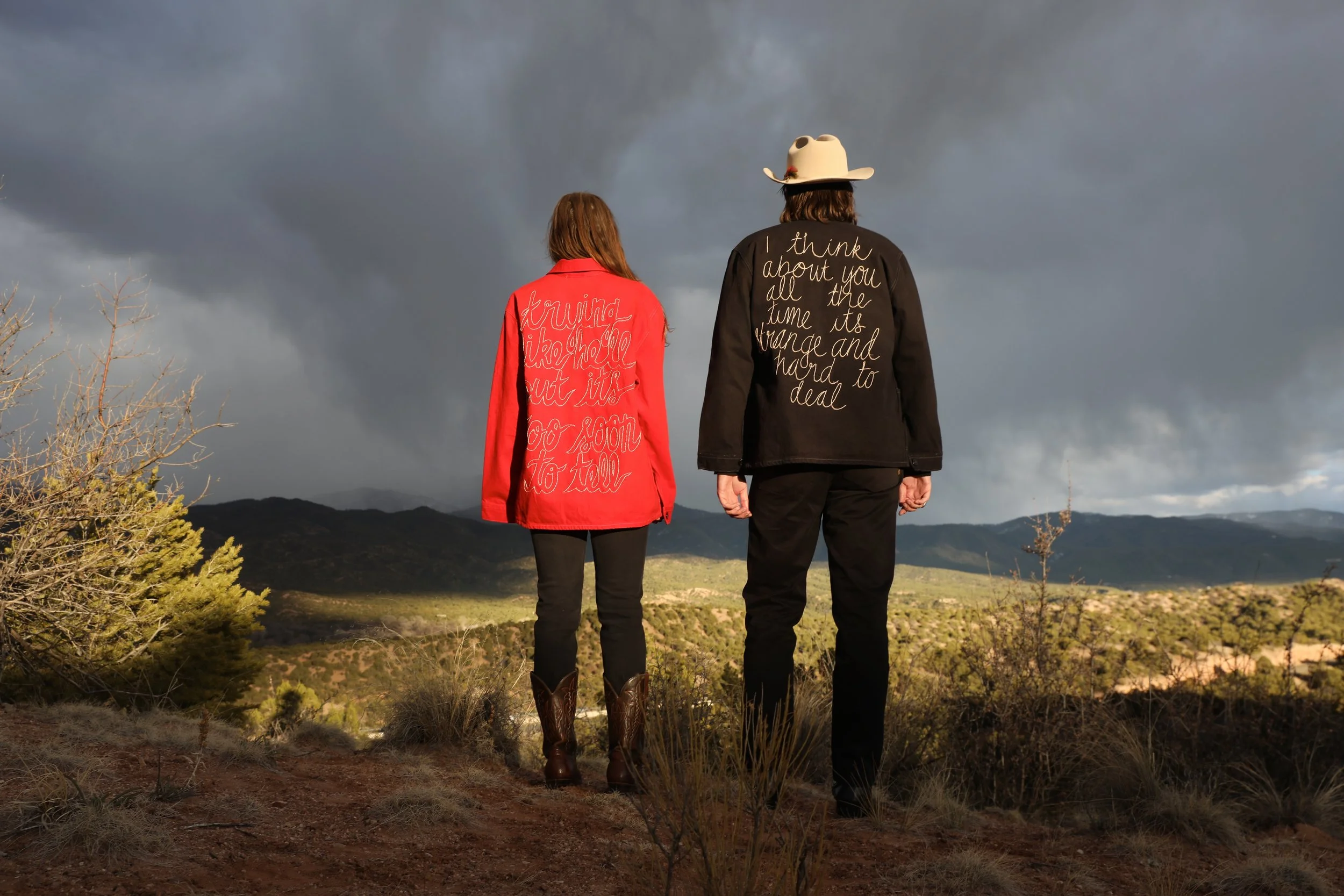 Two people are standing outdoors on a landscape with mountains in the background and dark stormy clouds overhead. One wears a red jacket with embroidered text on the back, and the other wears a black jacket with embroidered text on the back and a whi