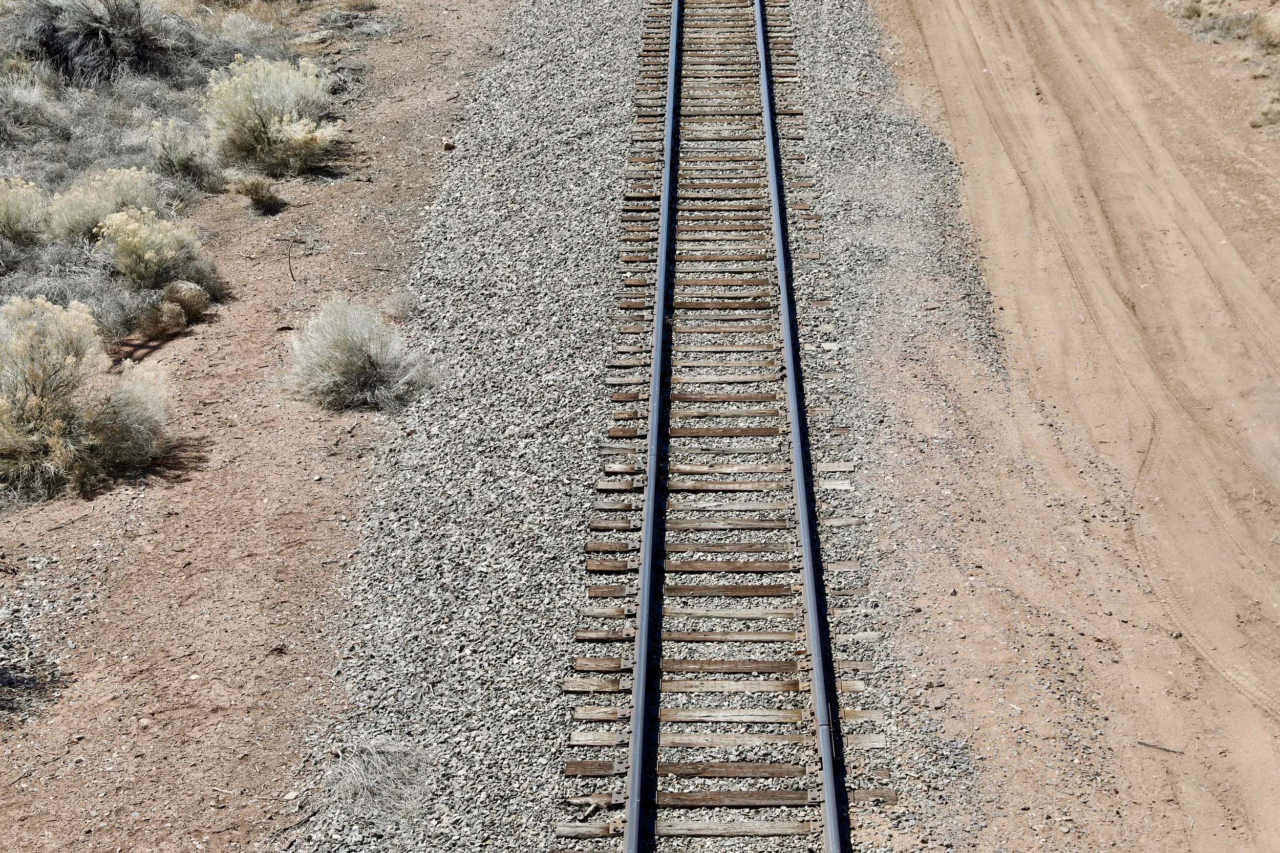 A straight railway track running through a desert landscape with sparse shrubs on one side and a dirt road on the other.