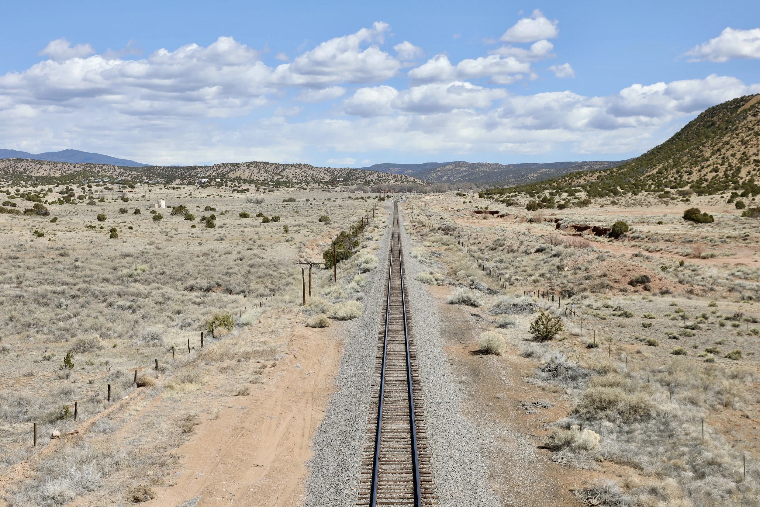 A straight railroad track extending through a dry, desert landscape with sparse vegetation and hills under a partly cloudy sky.