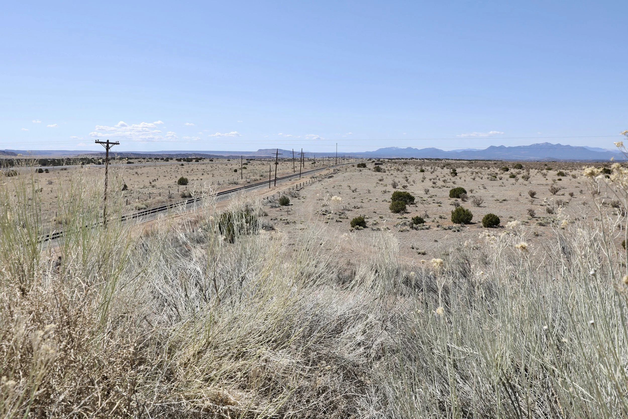 A desert landscape with sparse vegetation, power lines running parallel to railway tracks, and mountains in the distance under a clear blue sky.