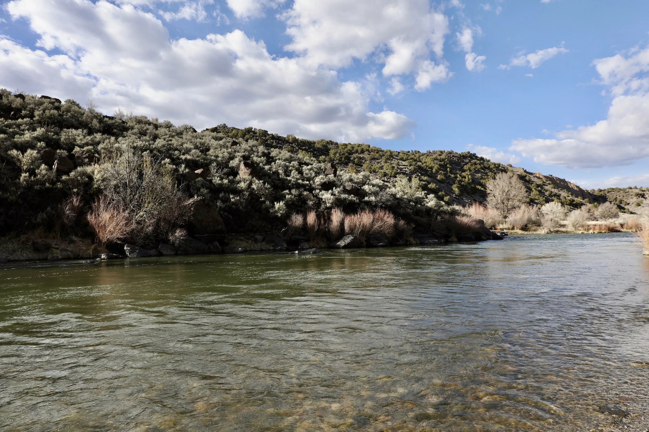 A river flowing through a hilly area with sparse trees and shrubs, under a partly cloudy sky.