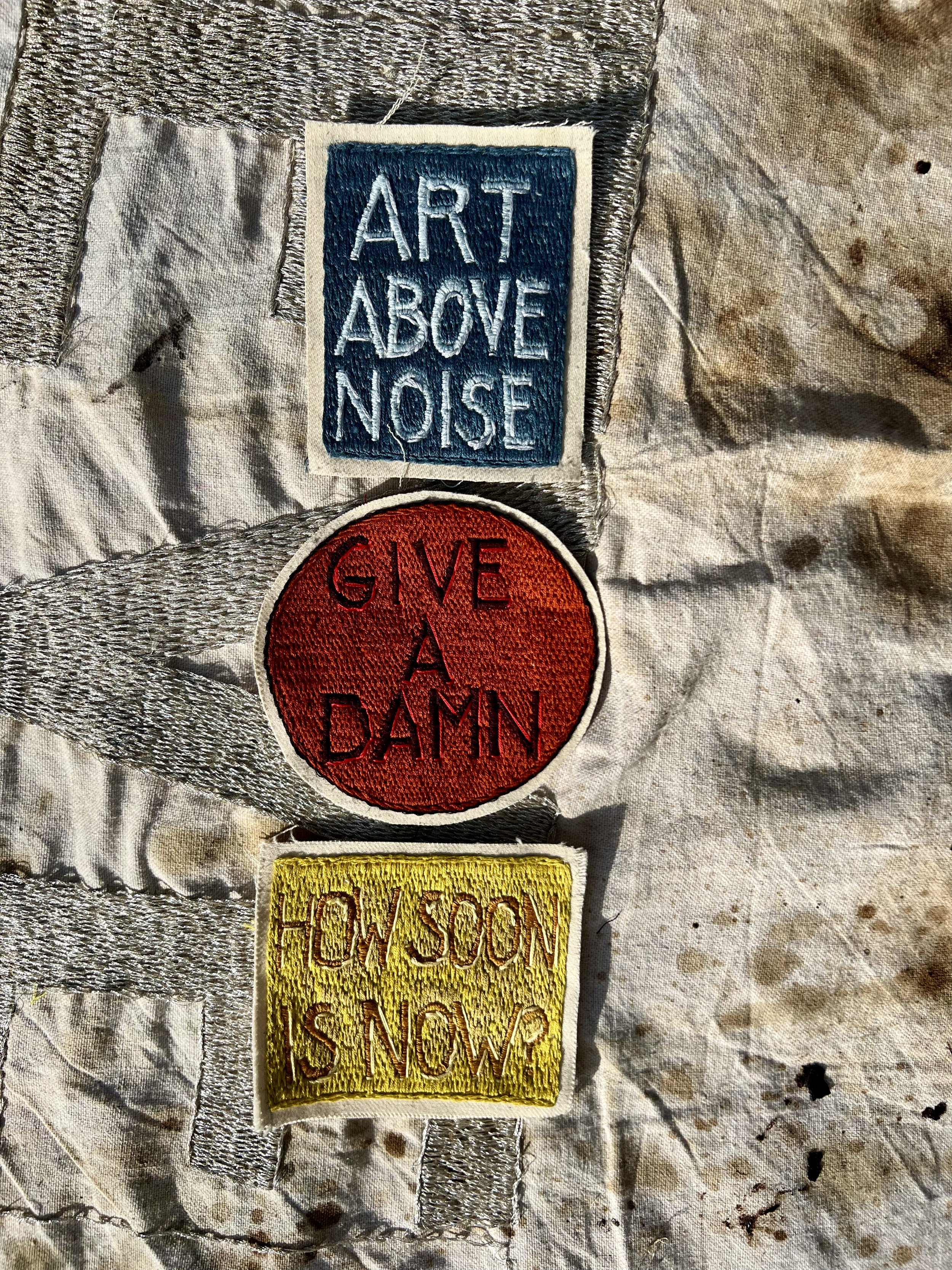 Close-up details of the Give a Damn PSA patch and Art Above Noise and How Soon Is Now hand-embroidered textile works styled on a canvas tote bag.