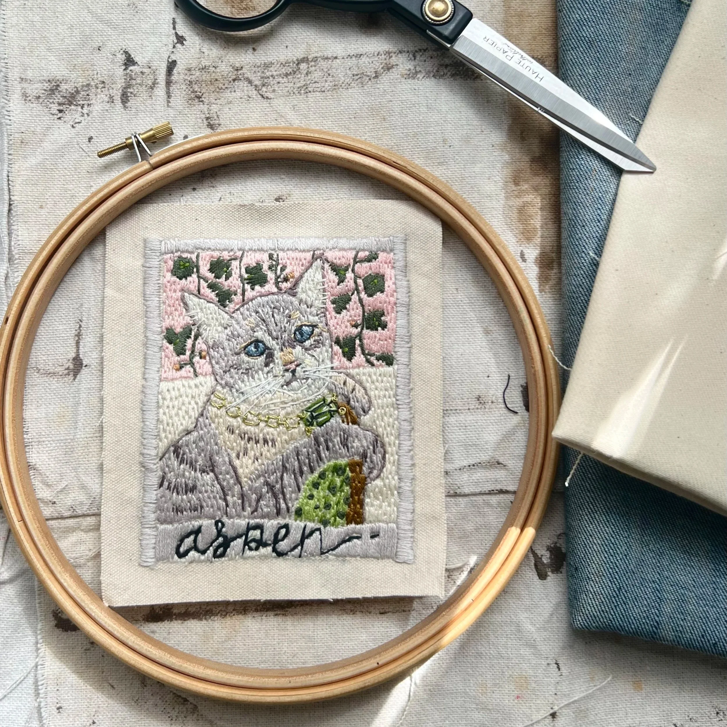 a hand embroidered custom portrait of aspen the cat. Set against a hand stitched victorian wall paper and filled with soul and light.