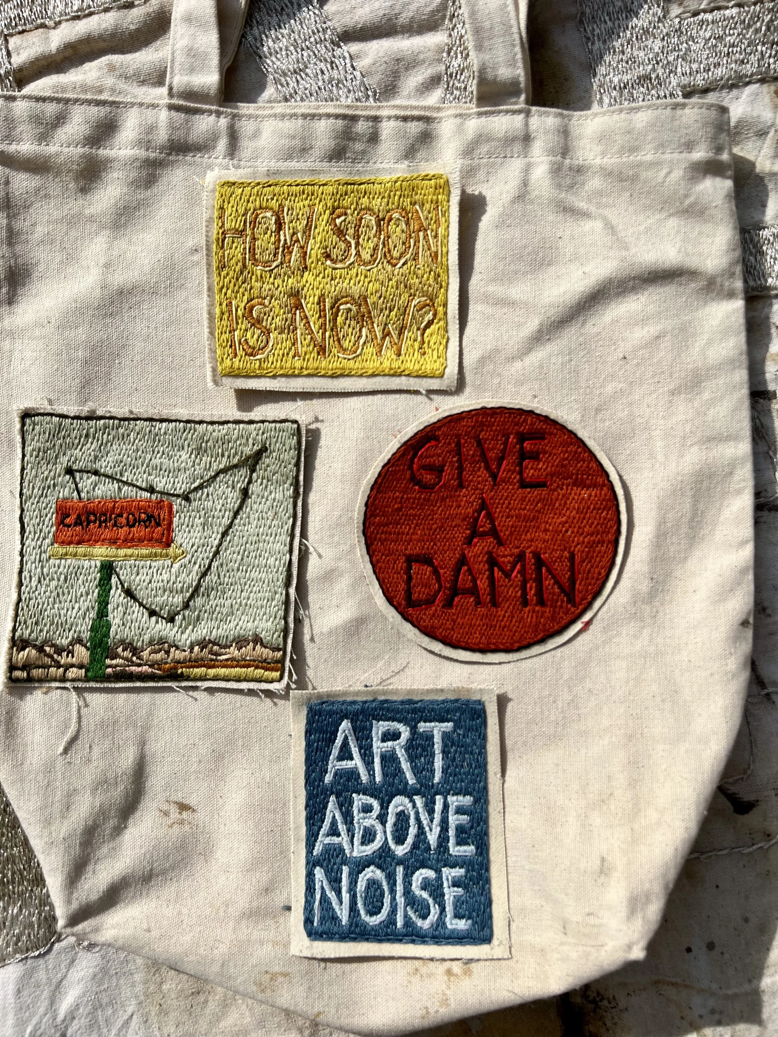 A close-up of hand-embroidered give a damn and art above noise patches stitched onto a canvas tote bag. Modern protest art, aclu support patch, artisan embroidery.