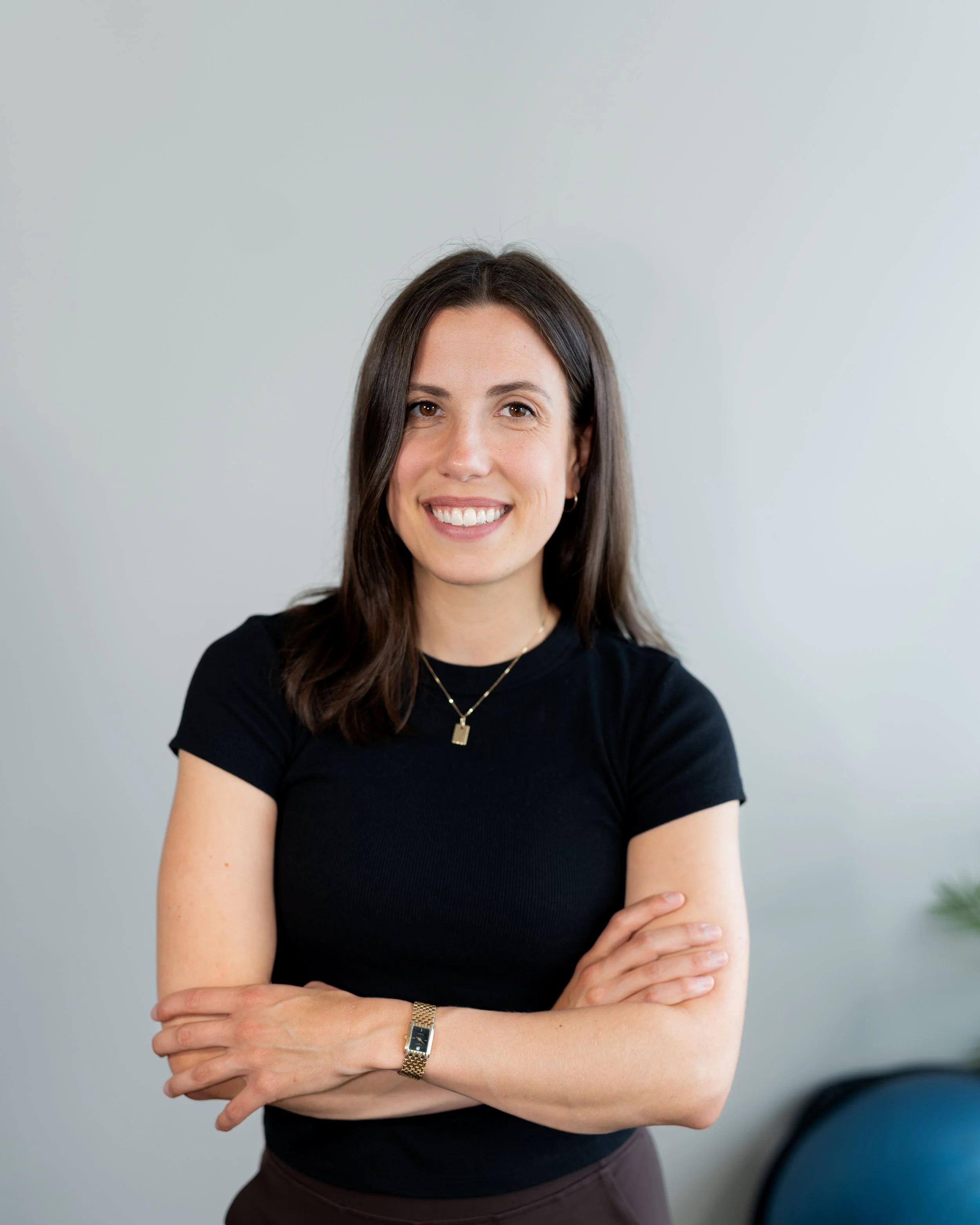 A woman with shoulder-length brown hair, wearing a black t-shirt, gold watch, gold necklace, and earrings, smiling with arms crossed against a plain light background.