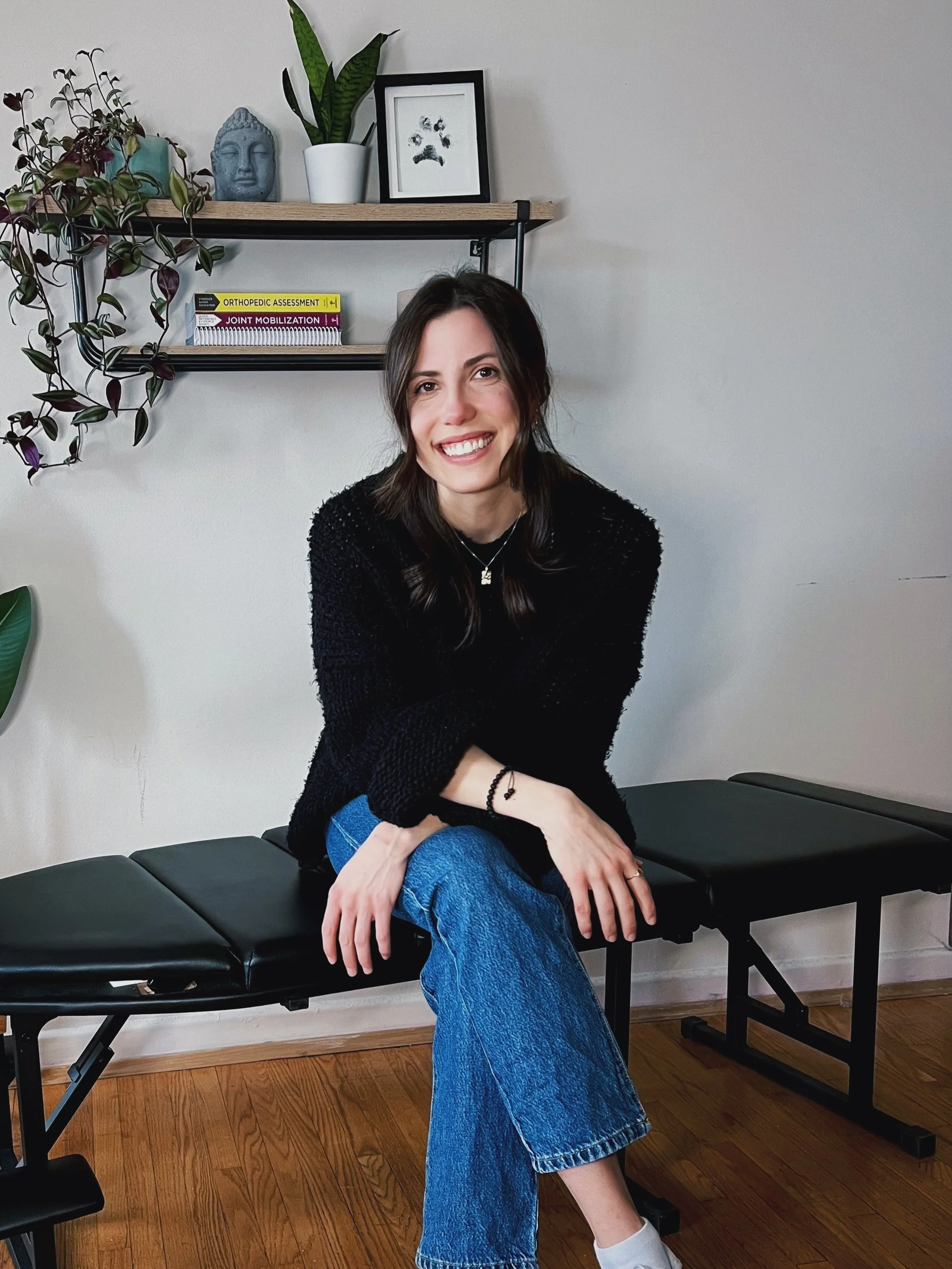 Smiling woman with dark hair sitting on a black chiropractic or medical examination table, wearing a black sweater and blue jeans, in a room with plants and books on a shelf.
