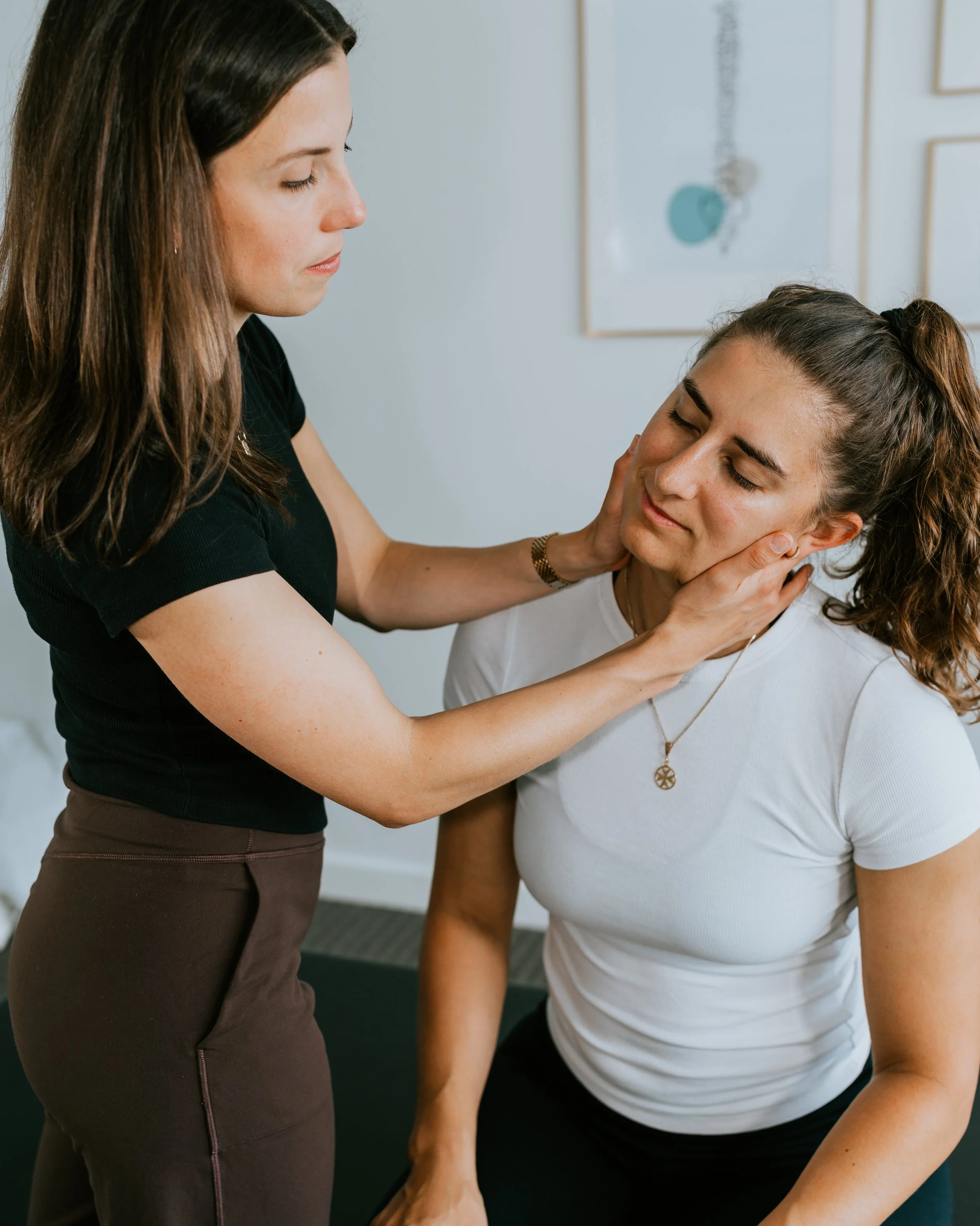 A woman in a black shirt gently holds the face of another woman with brown hair, dressed in a white shirt, who is seated with her eyes closed, in a comforting gesture in a clinical or therapy setting.