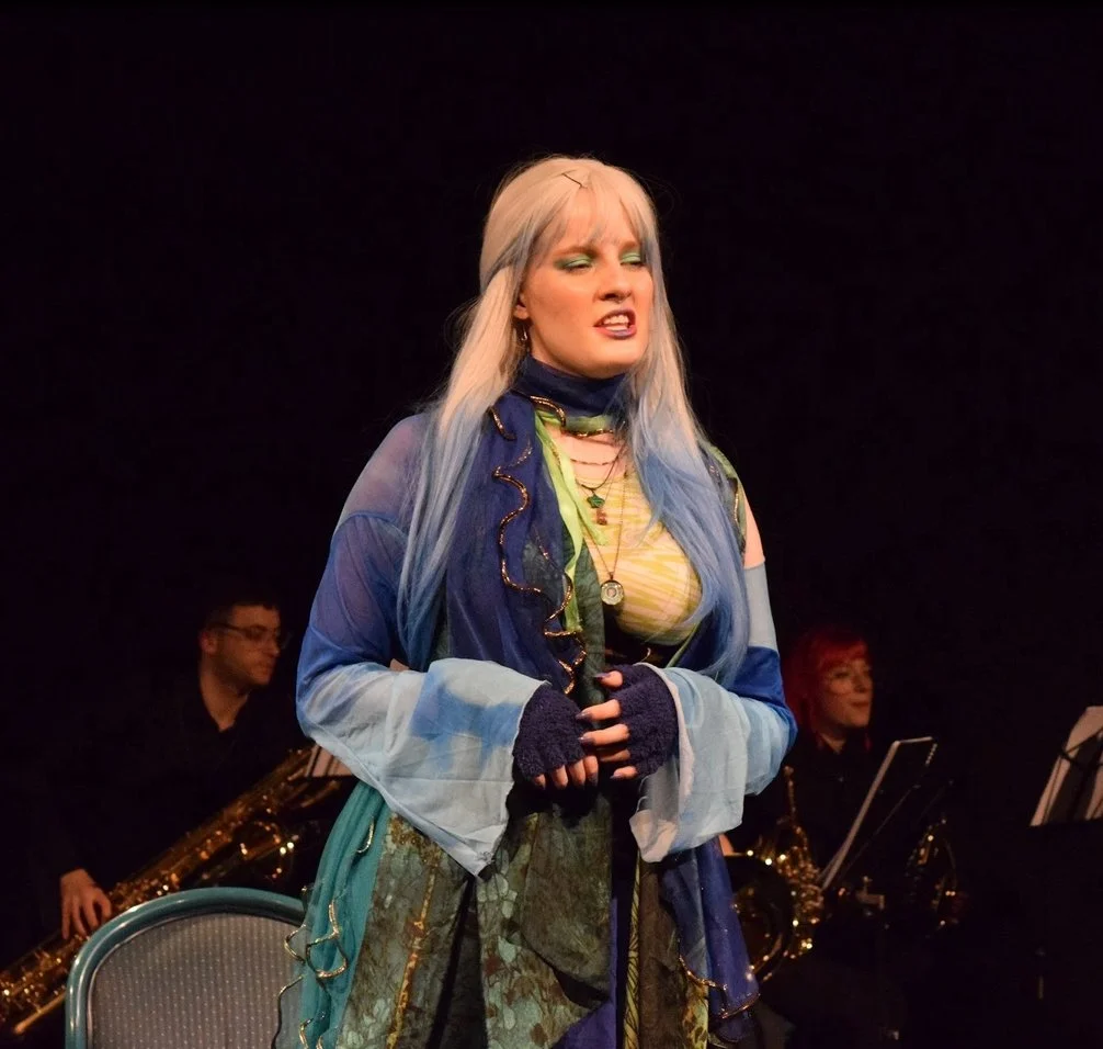 A female performer with long, multi-colored hair, wearing vibrant clothing and fingerless gloves, singing passionately on stage with musicians in the background.