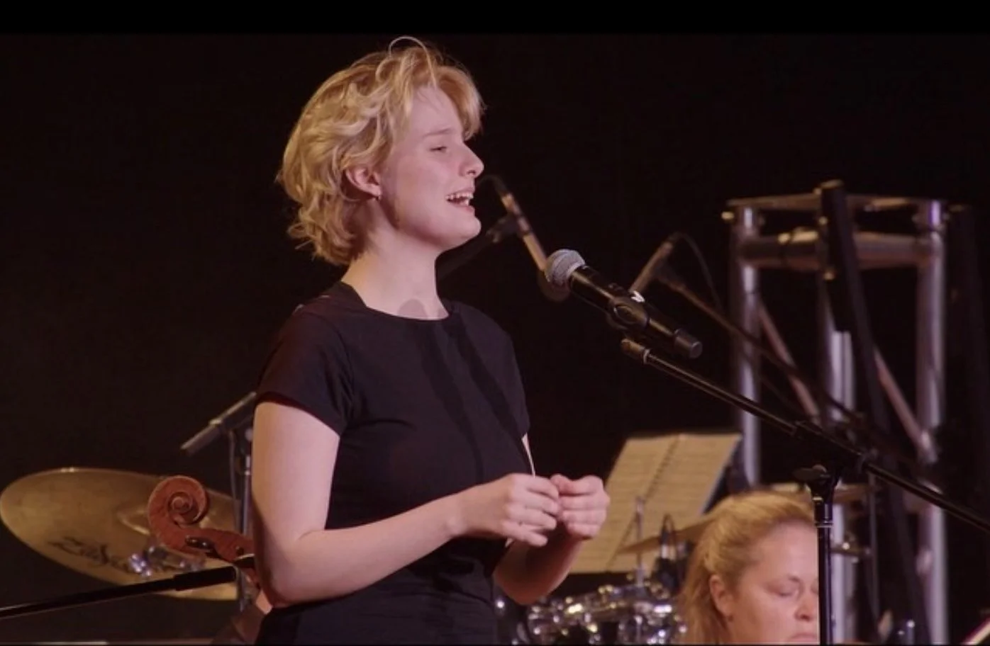 Young woman with curly blonde hair singing into a microphone during a performance, with a drum set and music stand in the background.